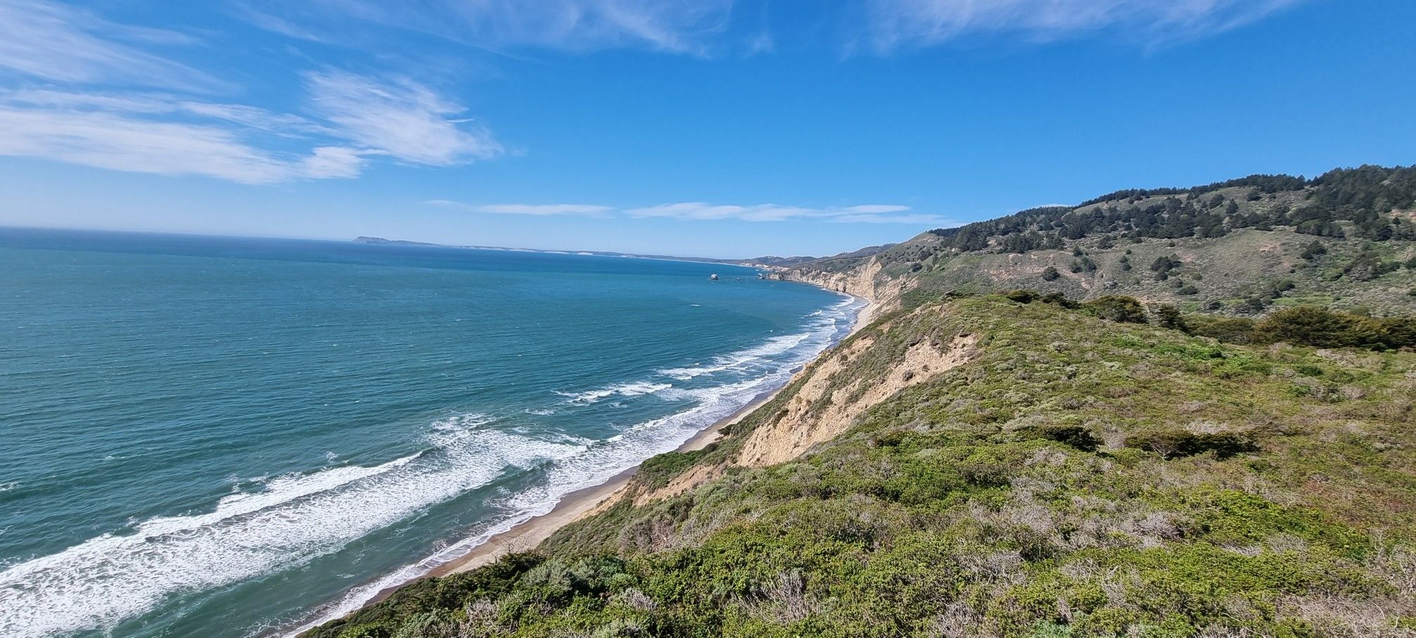 Panoramic view of a sunny coastline with green cliffs, blue ocean, and sandy beach. - Coast Trail and Lake Ranch Trail Loop