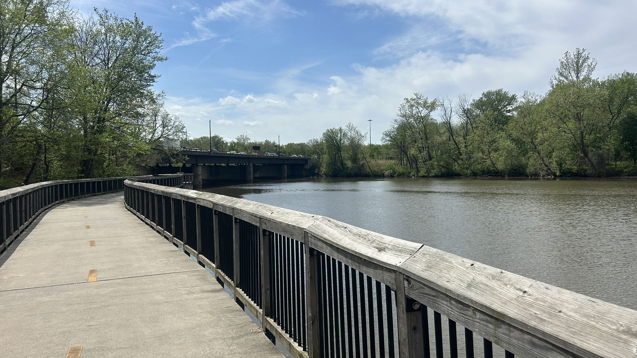Wooden boardwalk curves alongside a calm river with trees under a partly cloudy sky. - Anacostia Riverwalk Trail