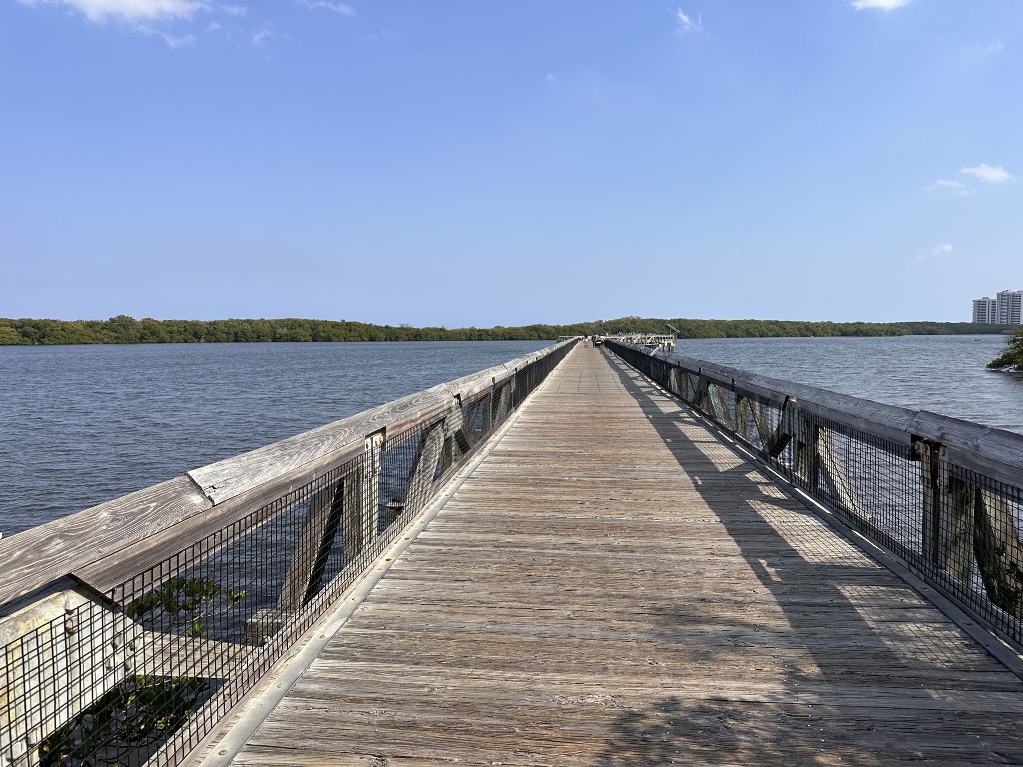 Wide wooden boardwalk stretches over calm water towards distant green shoreline. - John D. MacArthur Beach State Park Boardwalk