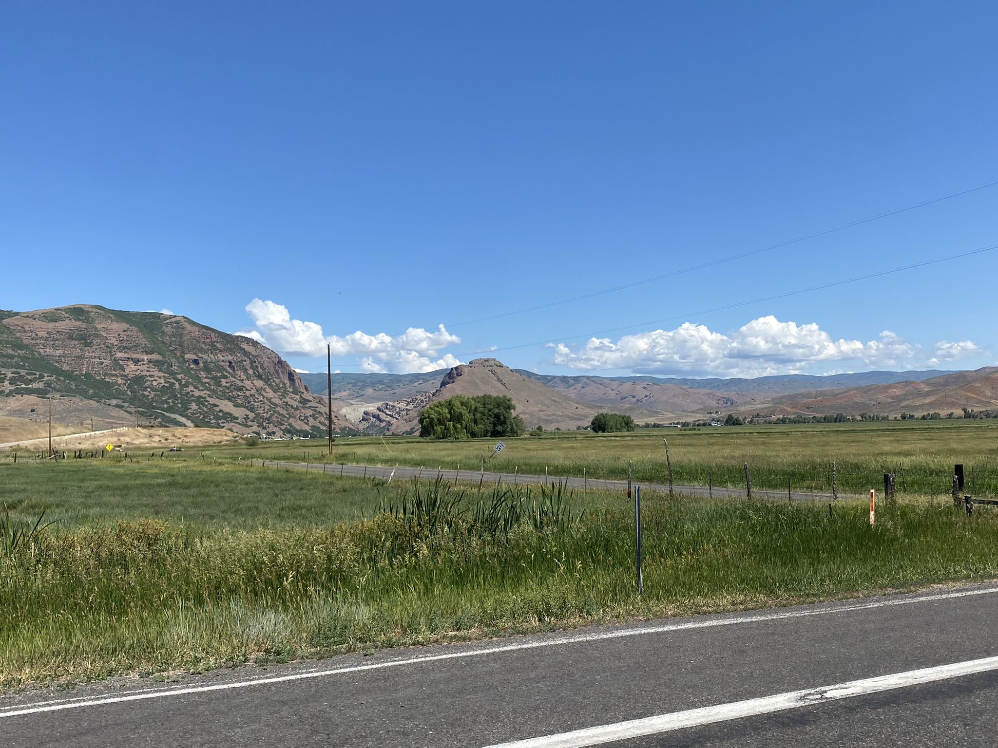 Road next to a vast green field with distant mountains under blue sky. - East Canyon Reservoir – Morgan Main Street loop from Henefer