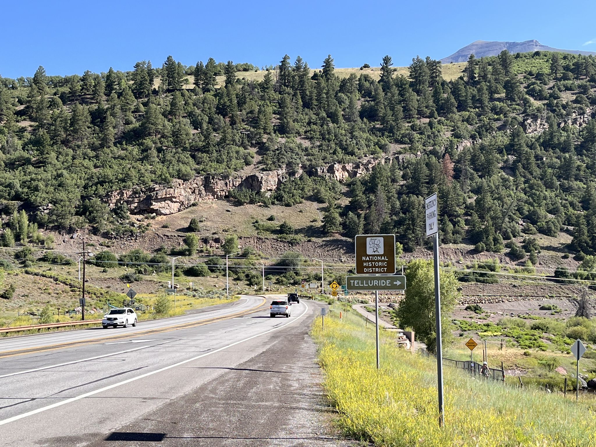 Cars drive on a highway next to a green, tree-covered mountain. - Telluride/Mountain Village loop
