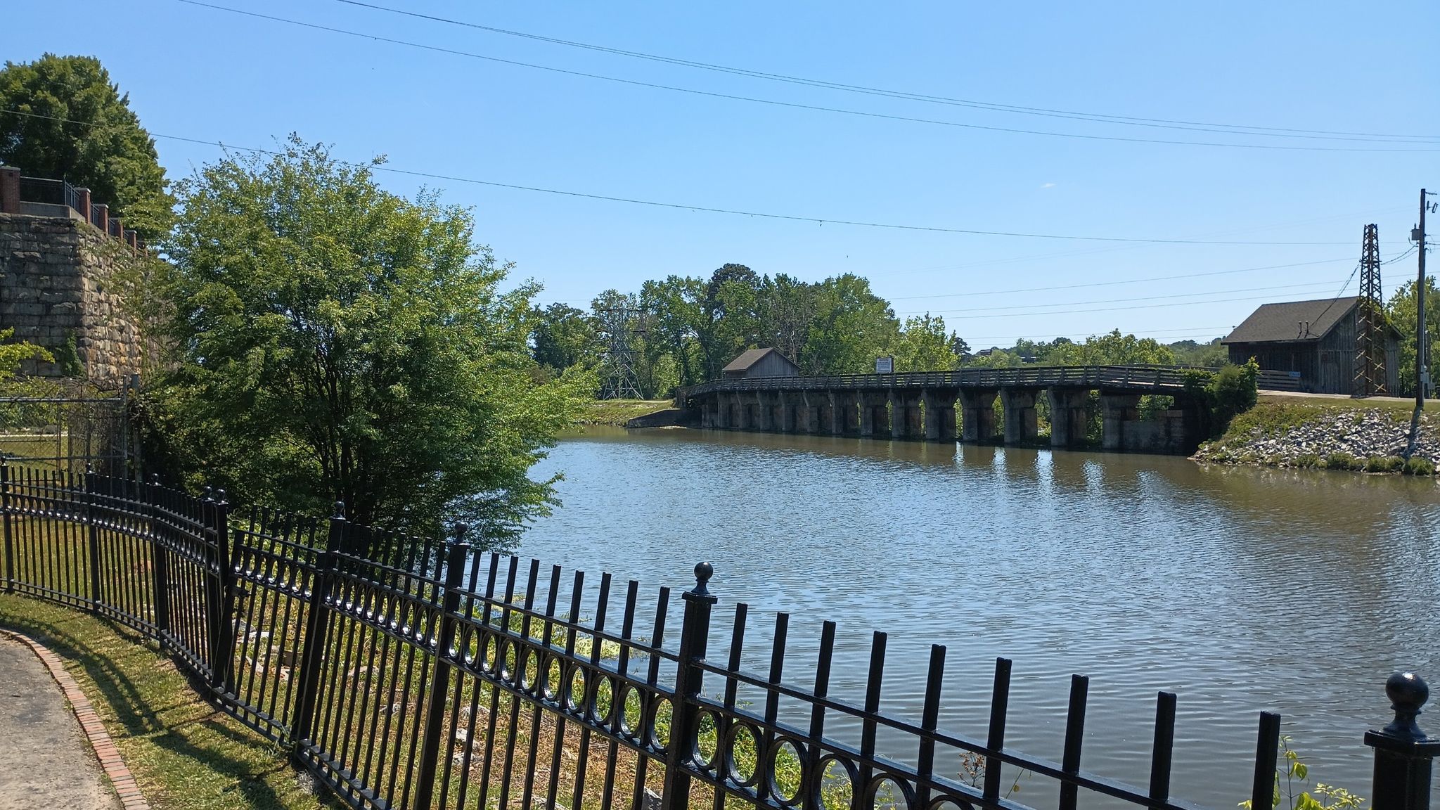 Calm water with a distant stone bridge and a black fence in sunny weather. - Riverfront Park Canal Greenway