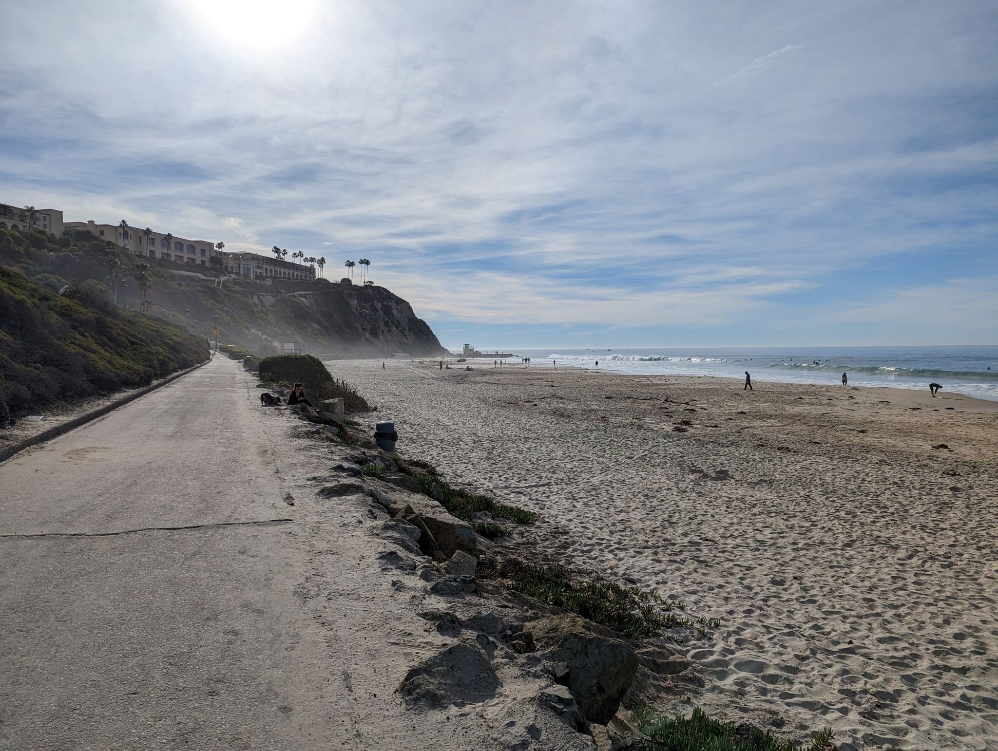 Sunny beach with ocean, paved path, and cliffside buildings. People walk on sand. - Salt Creek Trail