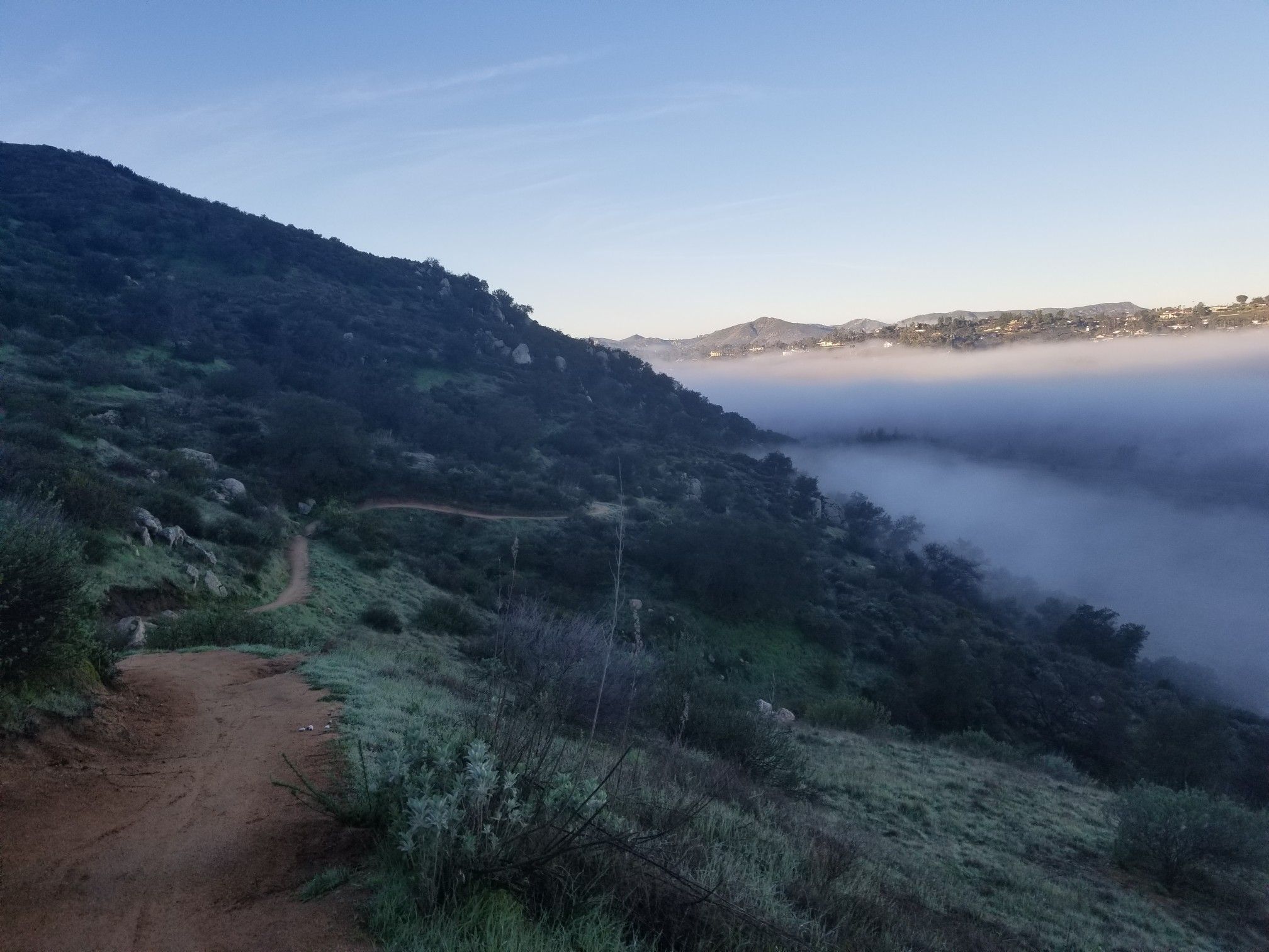 A dirt trail winds along a hillside overlooking a valley filled with fog. - Old Coach to Raptor Ridge
