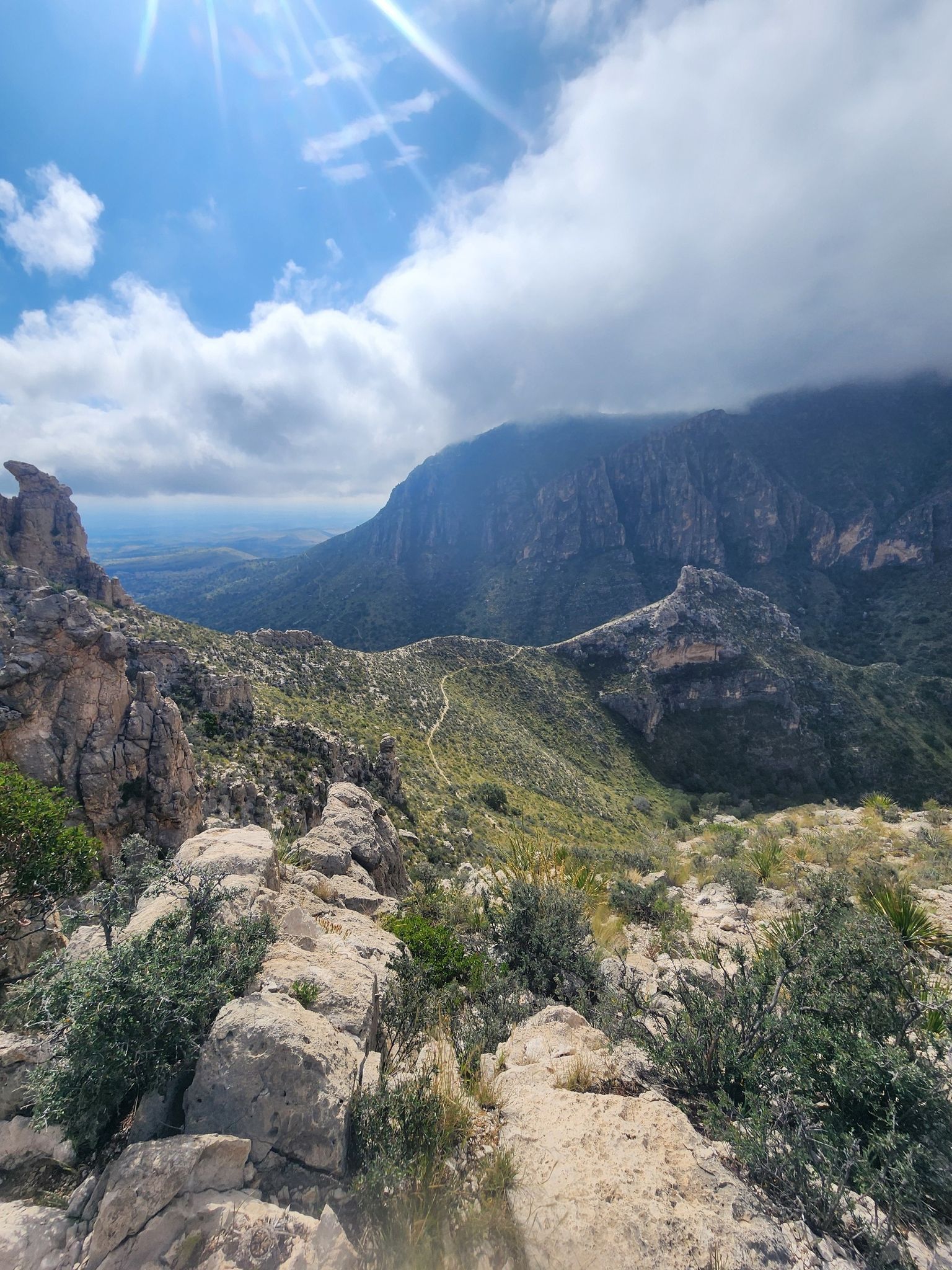 Sunlight illuminates rugged mountains, green valleys, and a winding path under cloudy skies. - The Bowl Loop
