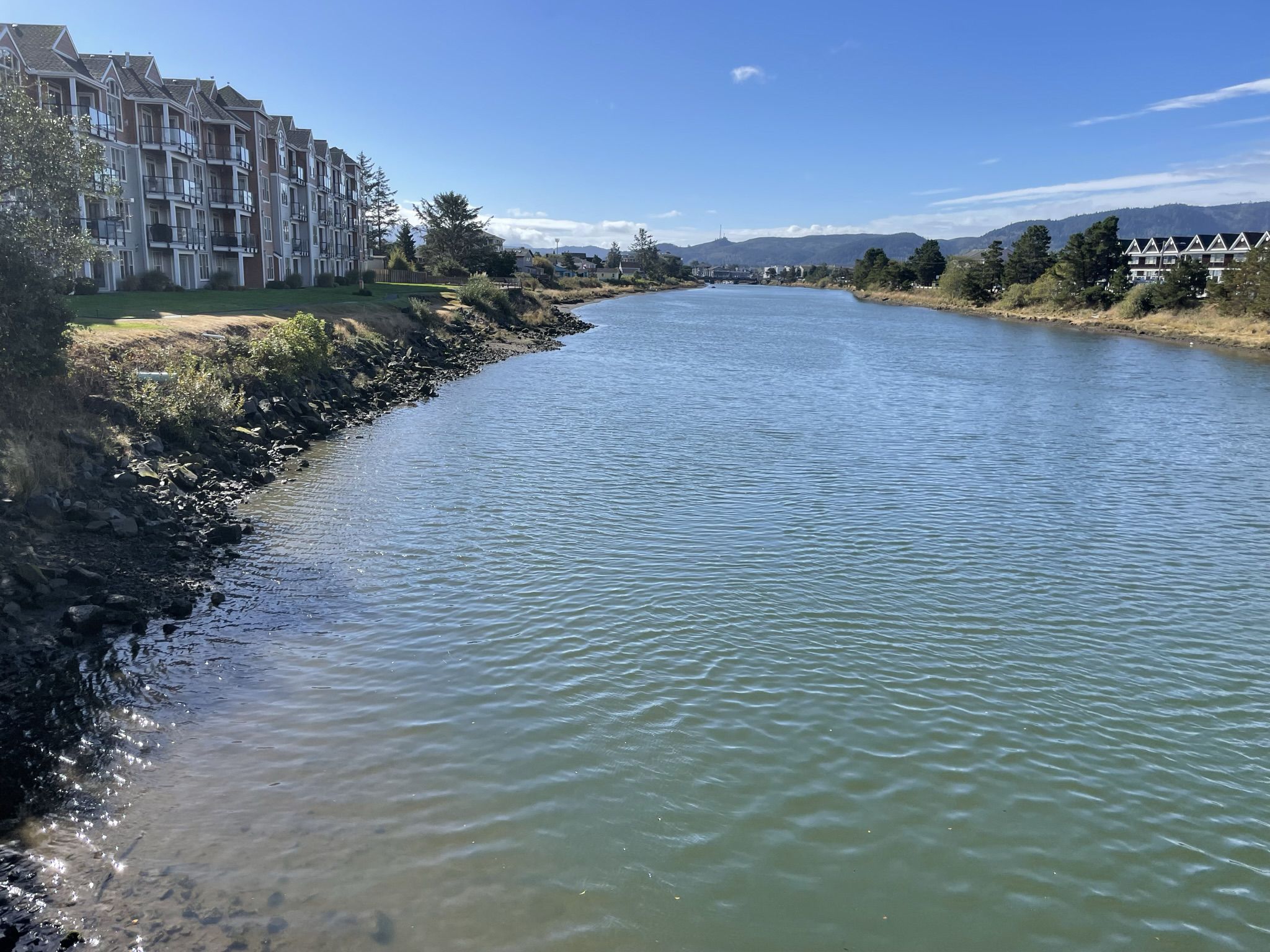 A wide river flows past buildings and distant hills under a clear sky. - TransAmerica Trail
