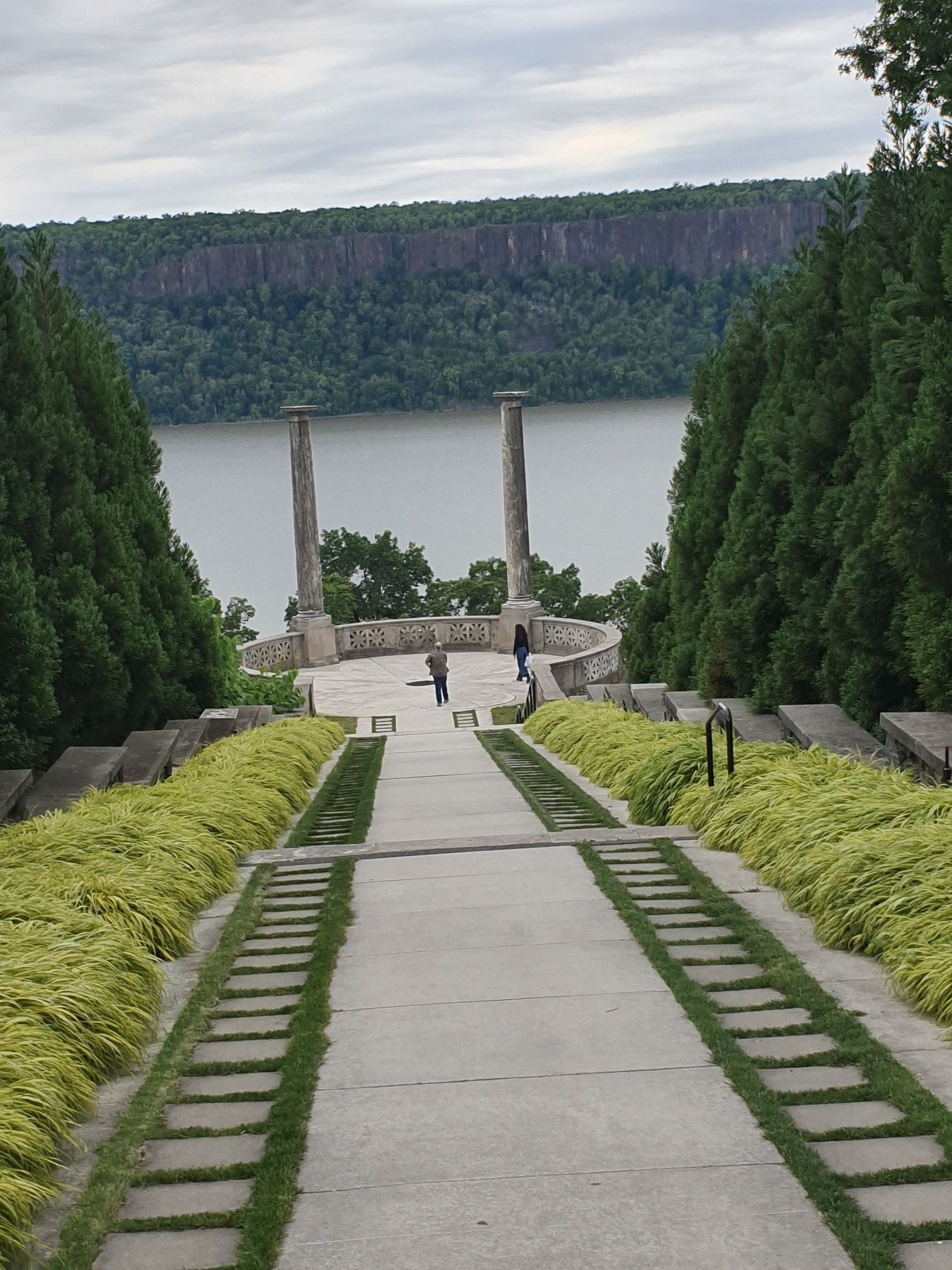 Long concrete path leading to stone columns by a river, people present. - Untermyer Park Loop