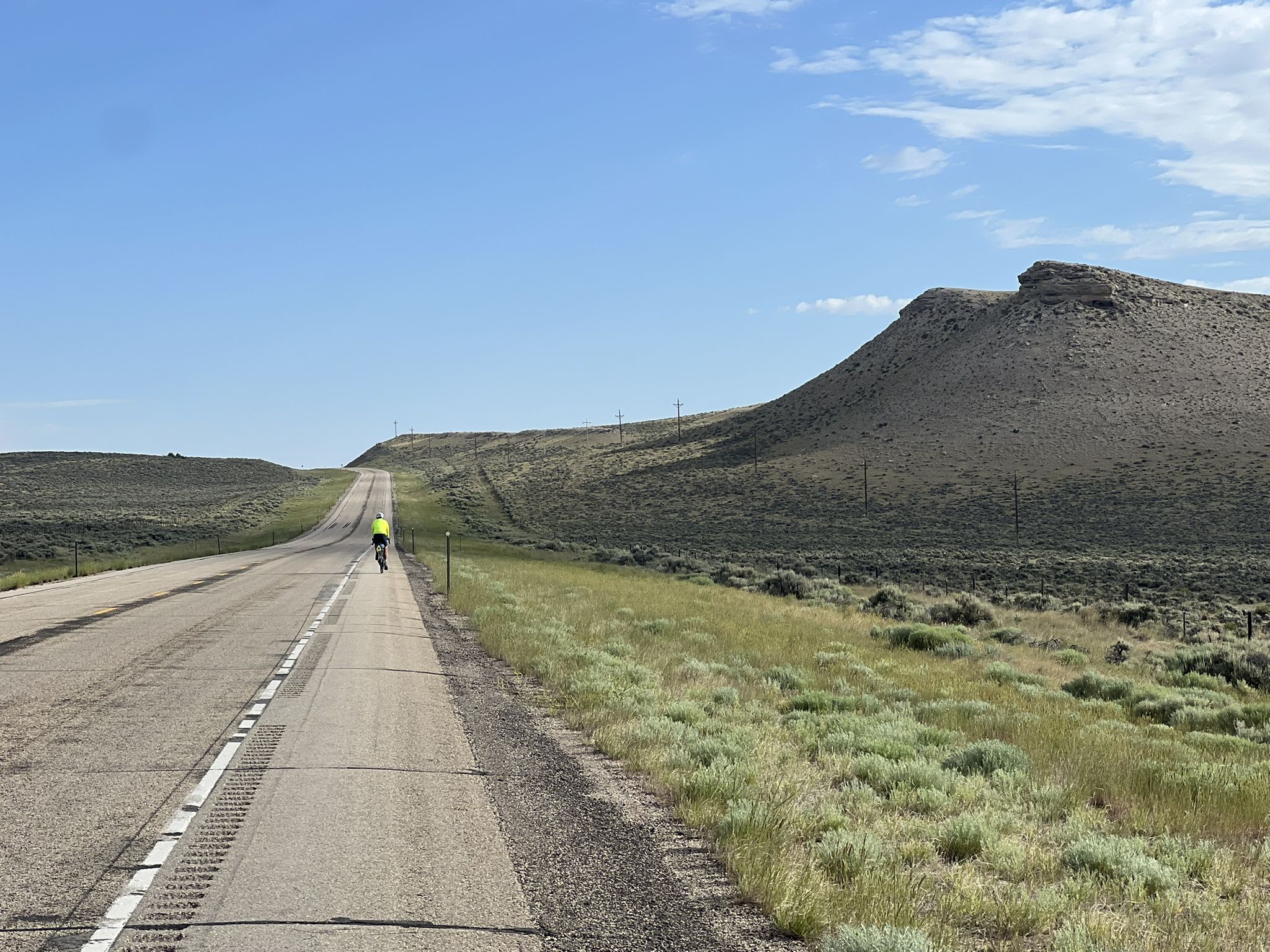 A cyclist rides on a long road beside a large, arid hill under a clear sky. - TransAmerica Trail: Saratoga to I-80 junction