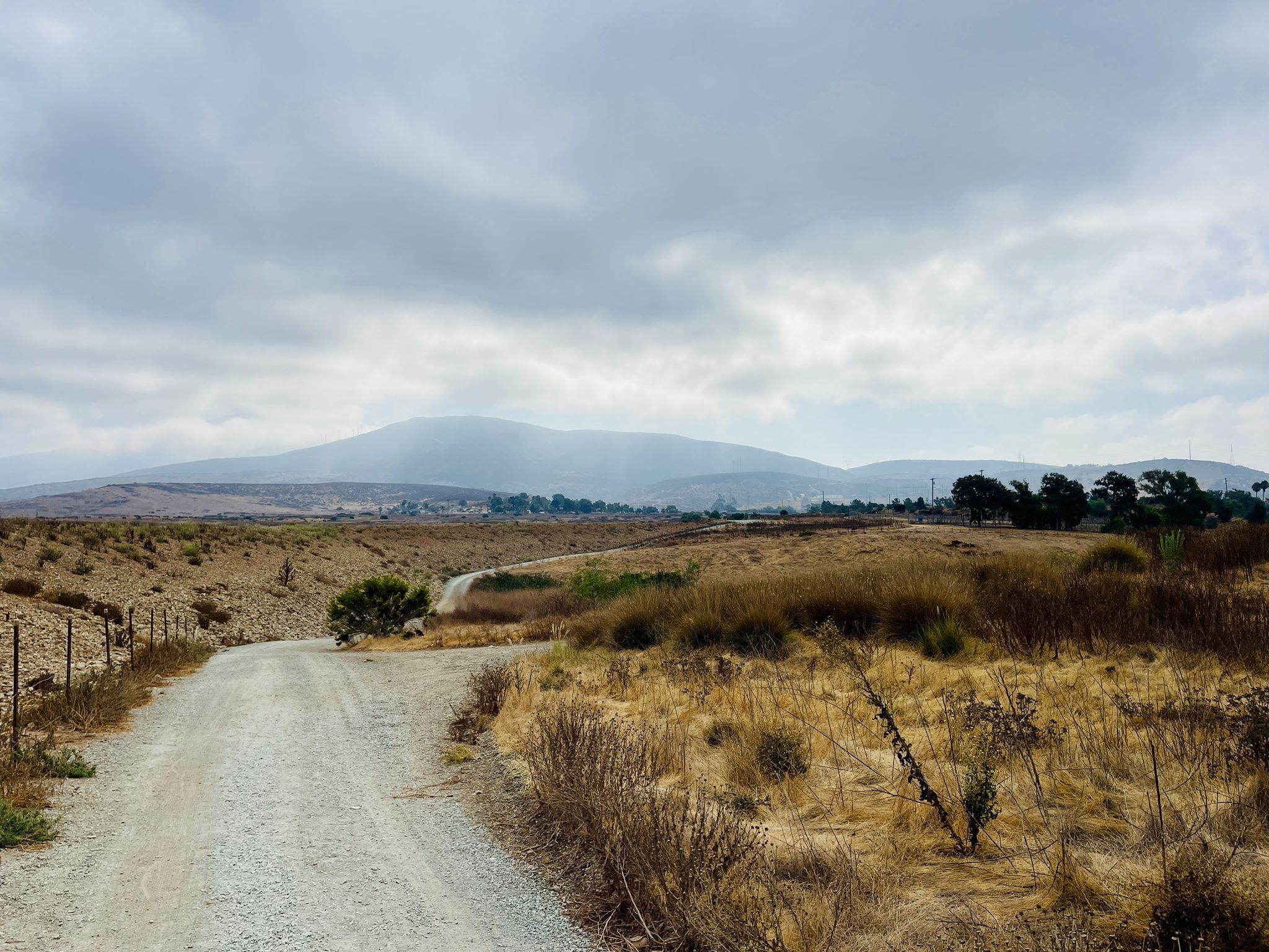 Dirt road winding through dry fields towards misty hills under cloudy sky. - Summit Park and Lake View Trail Loop