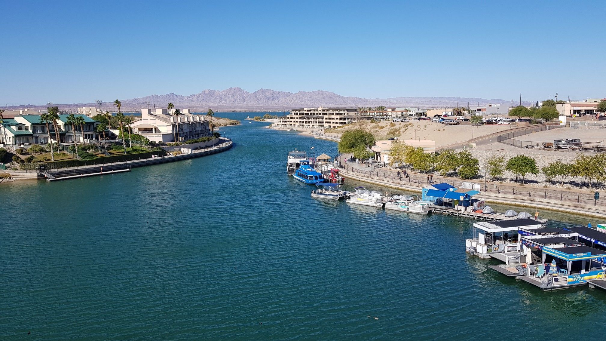 Sunny view of a waterway with boats, buildings, and distant mountains. - Mohave Sunset Trail