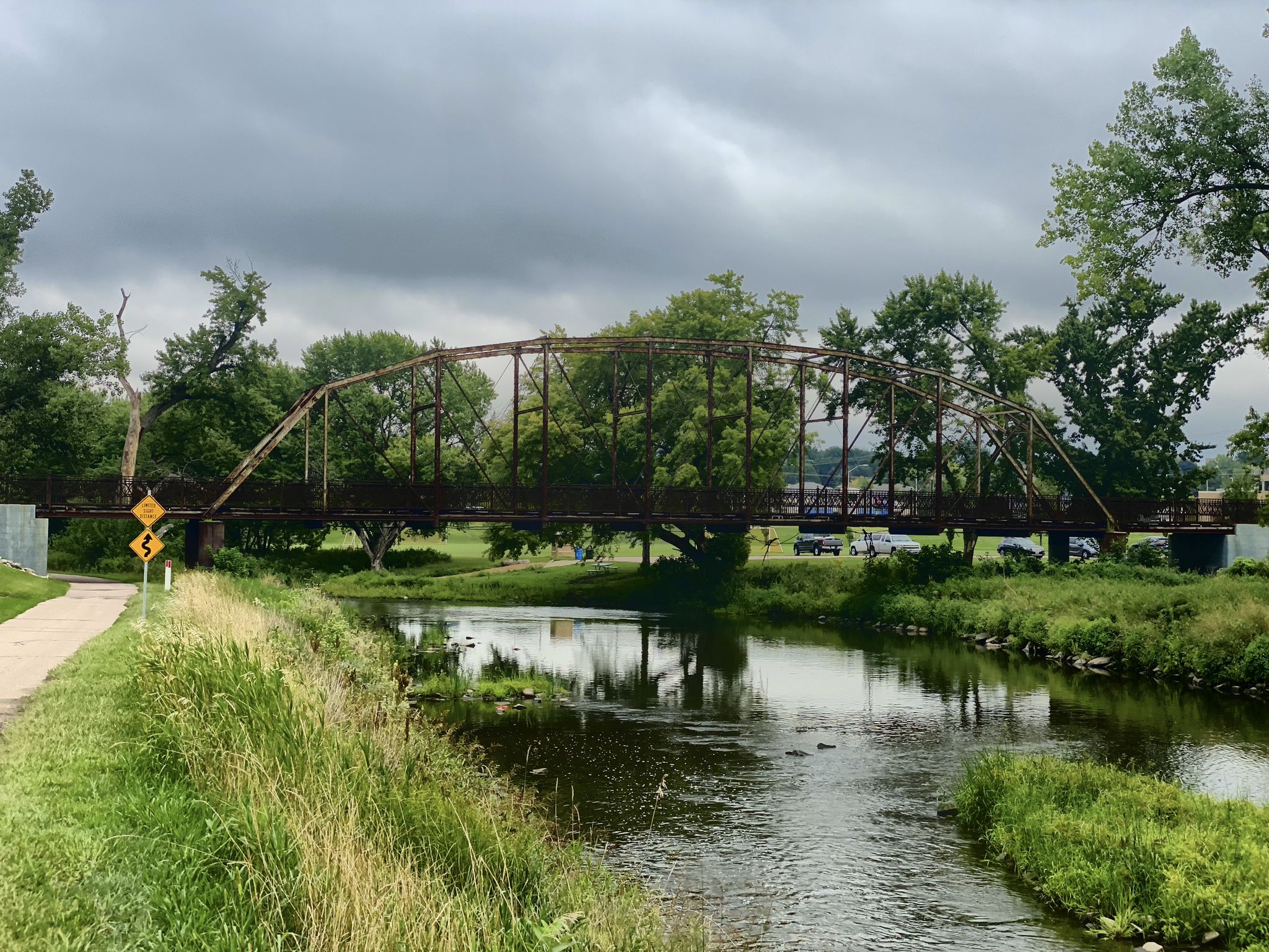 Old metal truss bridge over a river with grassy banks and trees. - Sertoma Park – Pedestrian Bridge loop from The Empire Mall