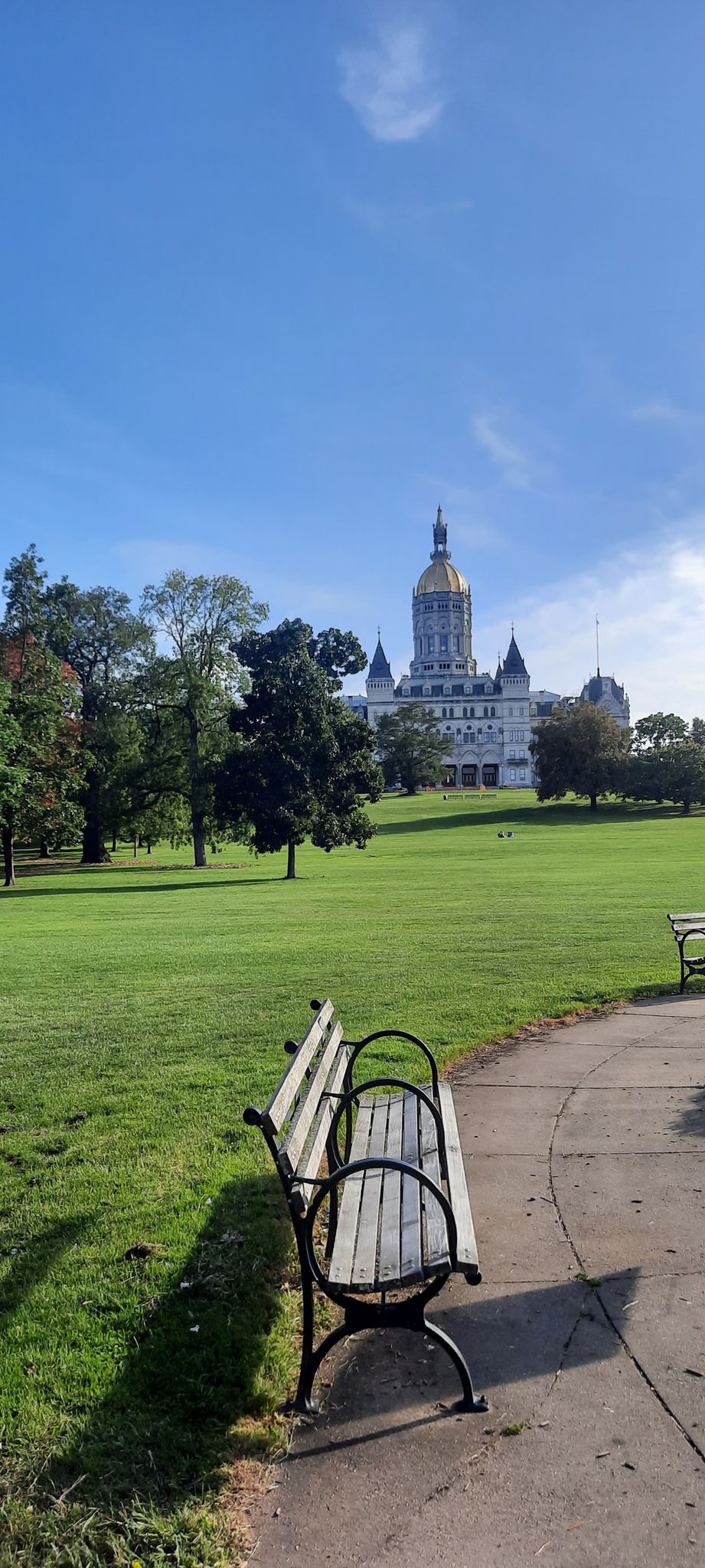 A park bench on a path with a large building and green lawn on a sunny day. - Bushnell Park – Elizabeth Park loop from Hartford