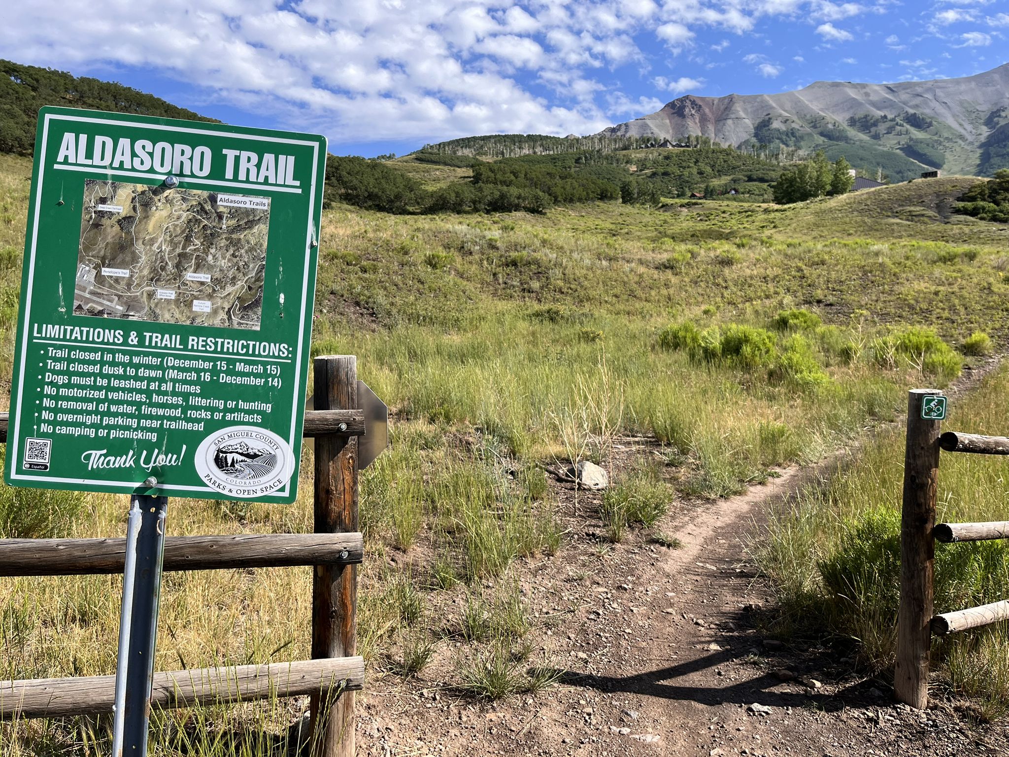 Green trailhead sign next to a dirt path leading into a grassy, sunny landscape. - Locals loop