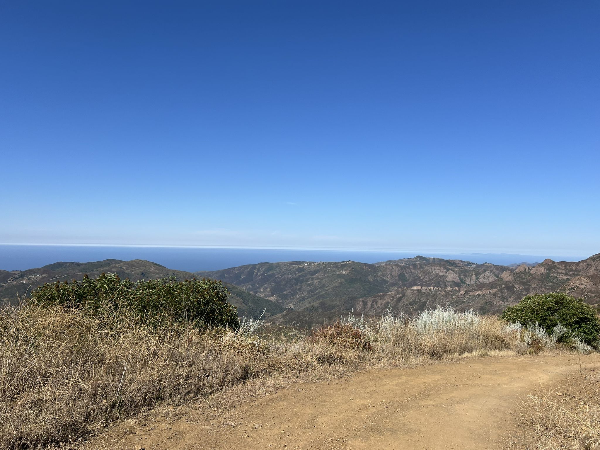 Dirt path overlooking vast green mountains and a distant blue ocean horizon. - MRCA Open Space – Long-distance hiking trail loop from Backbone Trail - Encinal to Mulholland