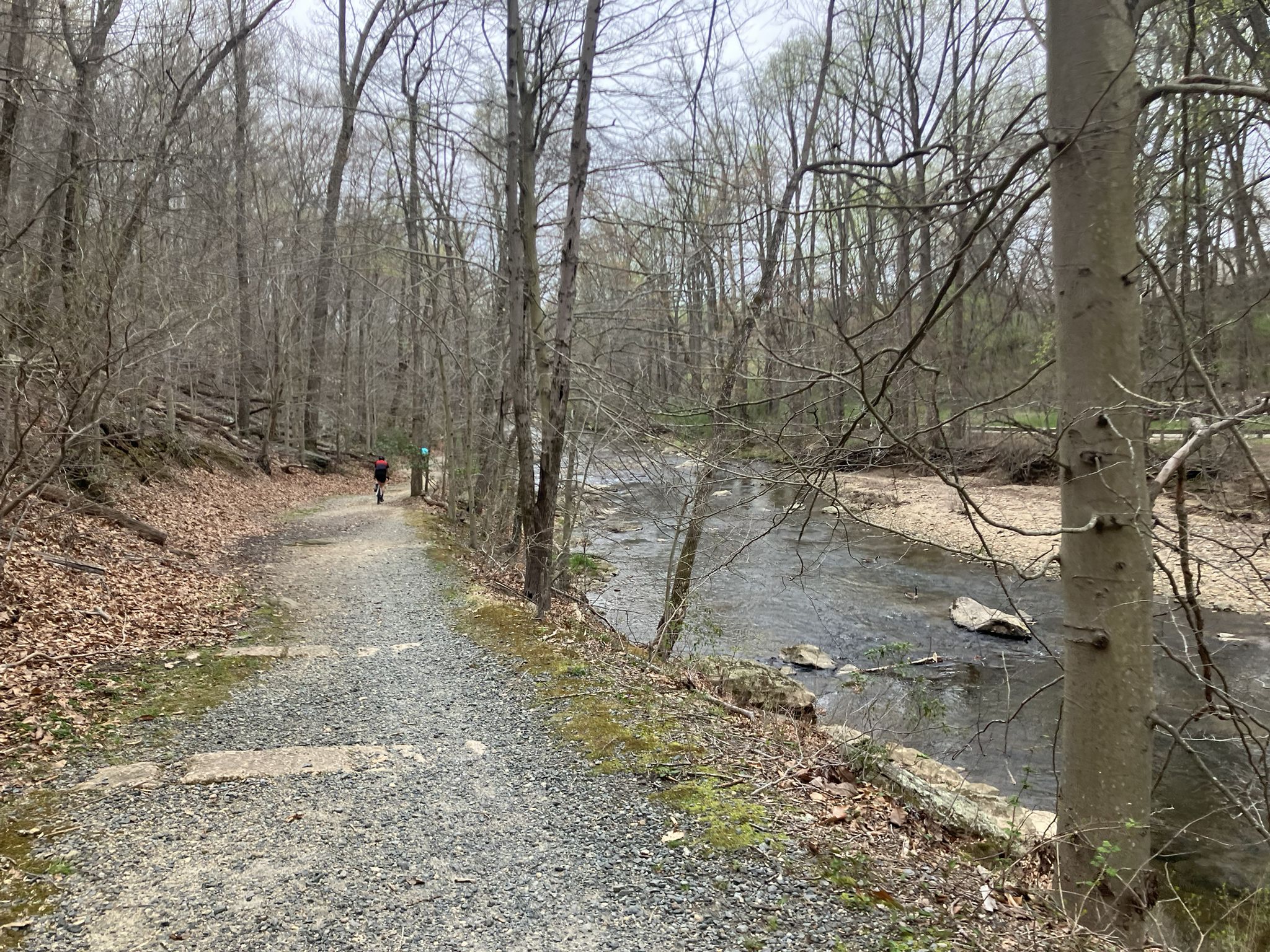 A person walks on a gravel path beside a rocky river with bare trees. - Fair Hill Trailhead – Alexander Ruins loop from Fair Hill