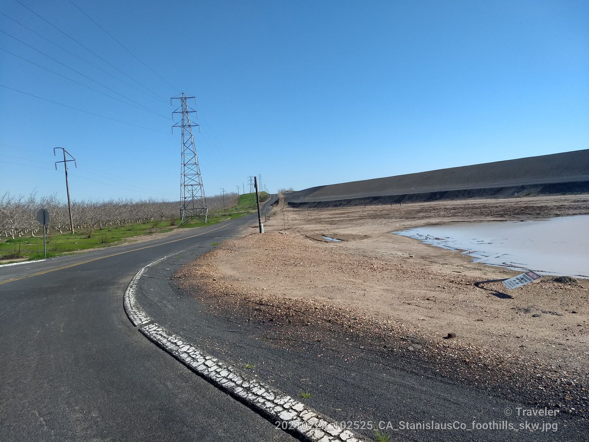 Winding road, power lines, and a dirt mound with a water puddle. - Woodward Reservoir dam 🌊 – Woodward Reservoir loop from Adela