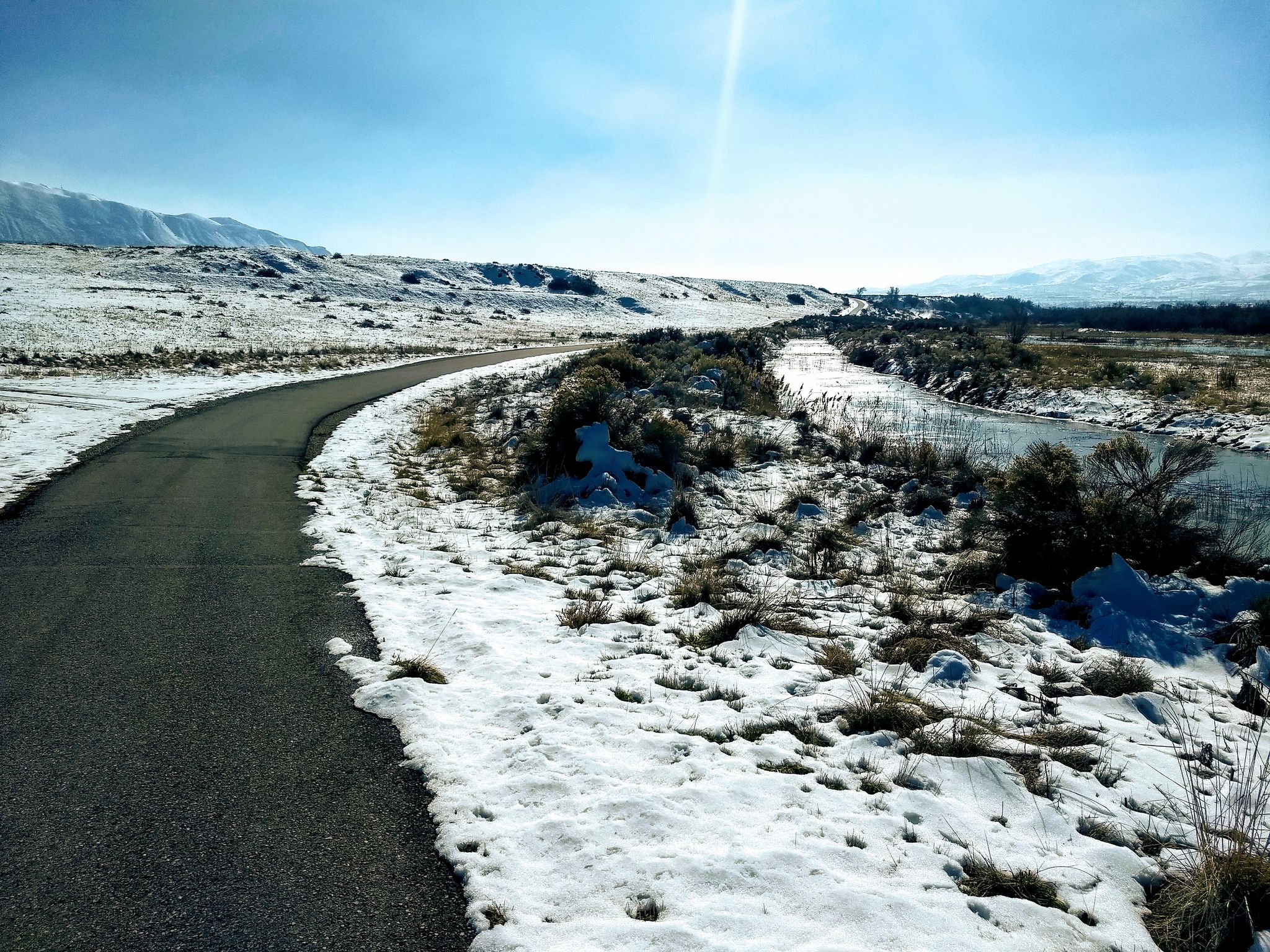 Paved road winding through a snowy, sunny landscape with a river. - Riverfront Parks – Riverfront Parks loop from Fashion Place West
