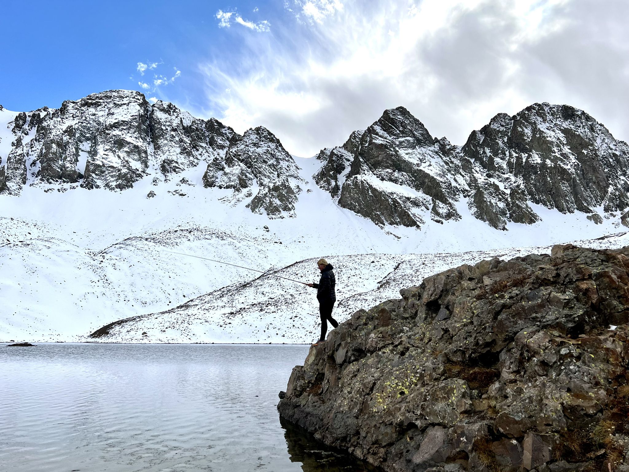 A person fishes from a rock beside an alpine lake with snowy mountains. - Cinnamon Pass