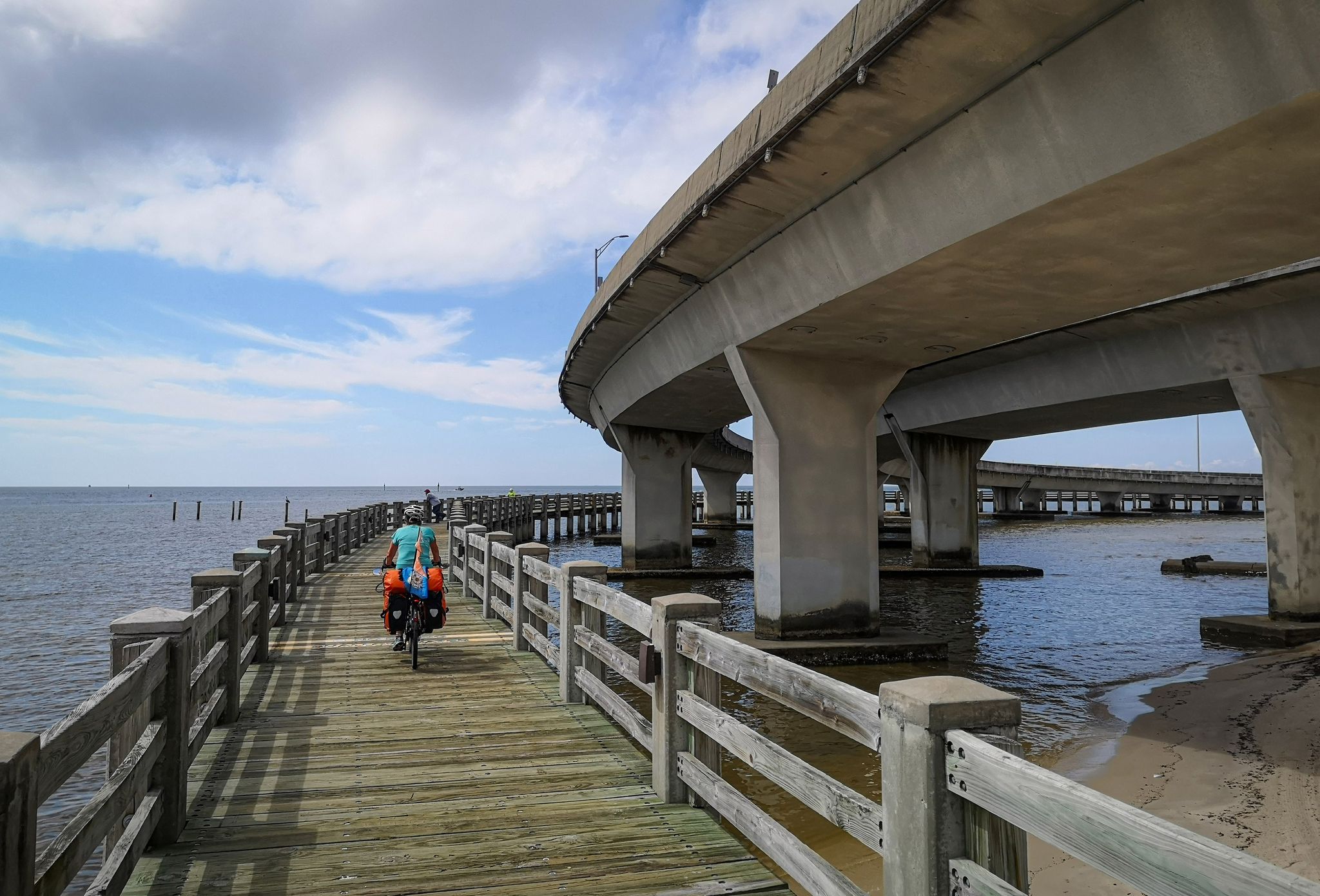 A cyclist on a wooden pier passing under a large concrete bridge over water. - Biloxi West Beach Boardwalk