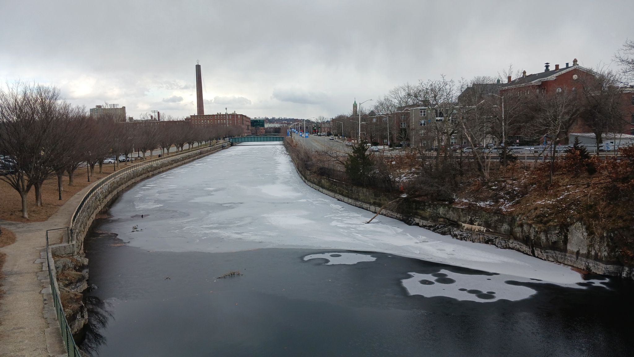 Overcast view of a partially frozen waterway lined with bare trees and buildings. - Lowell Riverwalk