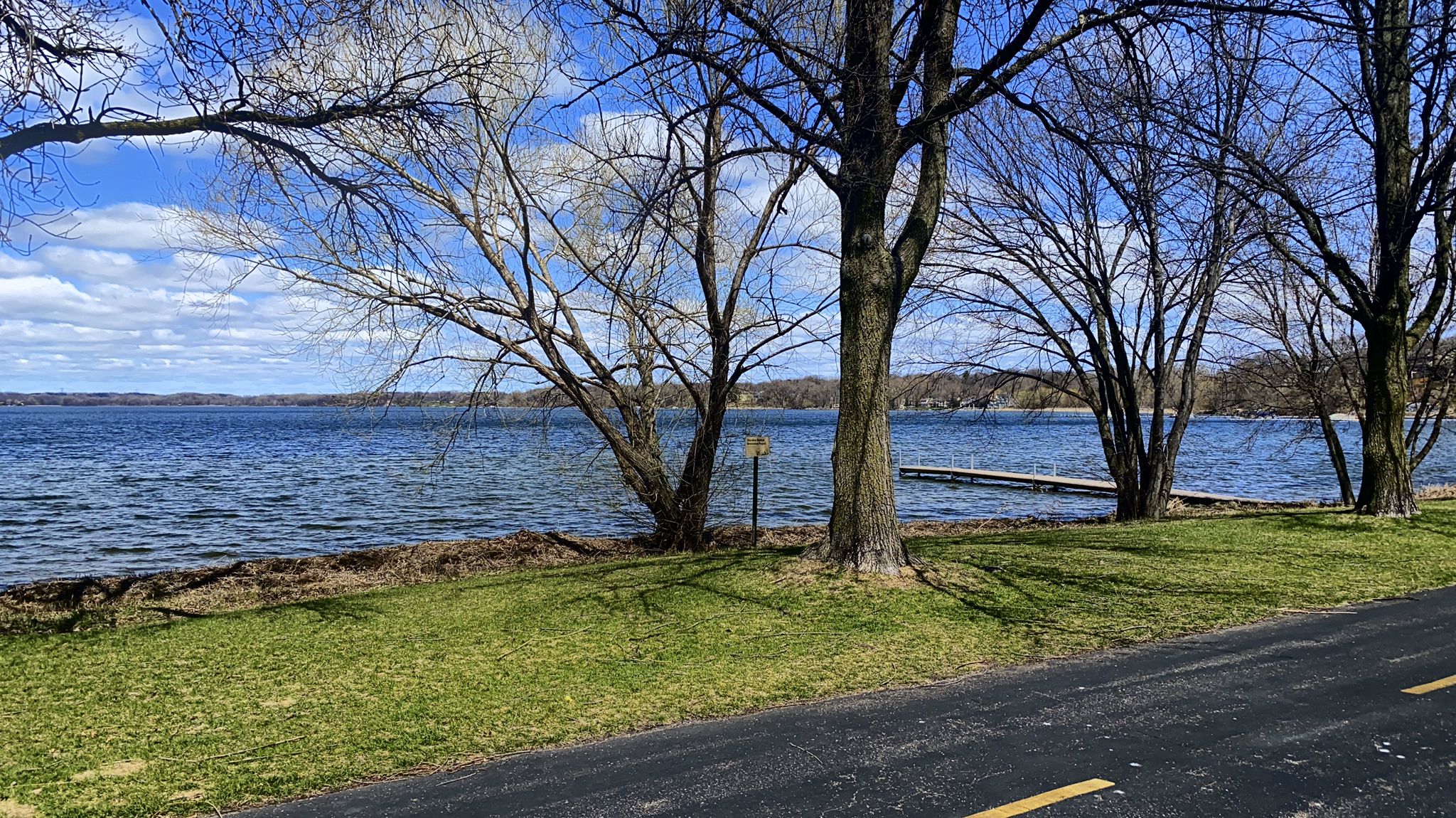 Bare trees line a lake shore next to a road on a sunny day. - Luce Line Brewing – Boardwalk loop from Medicine Lake