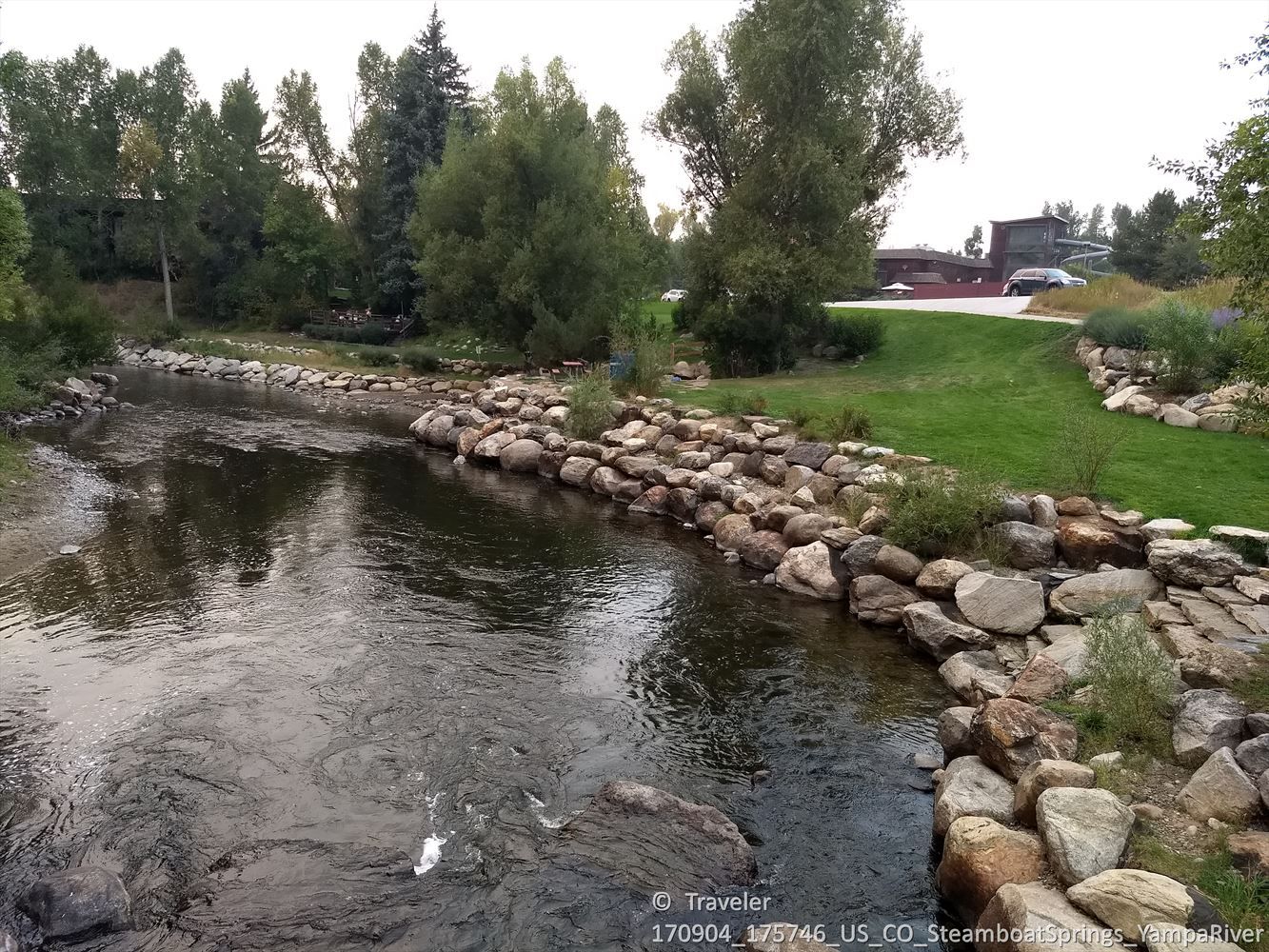 River with rocky banks and green grass, bordered by trees and buildings. - Yampa River Core Trail