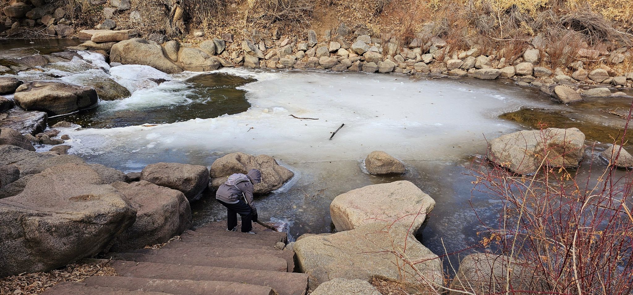 A person stands on steps by an icy river with large rocks. - Boulder Creek Path