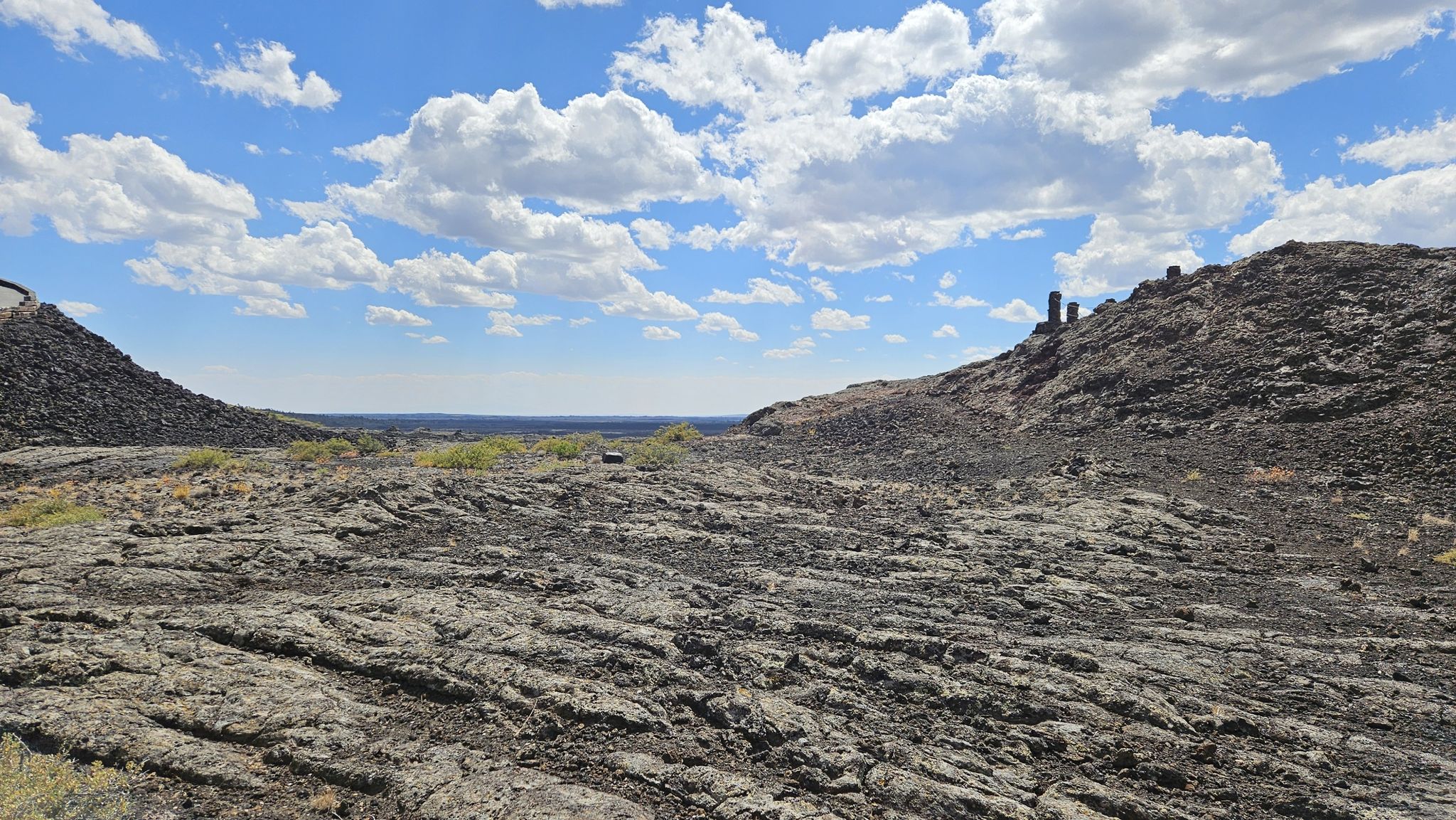 Wide view of dark, textured volcanic rock under a bright blue sky with clouds. - Snow Cone Trail & Spatter Cones Trail