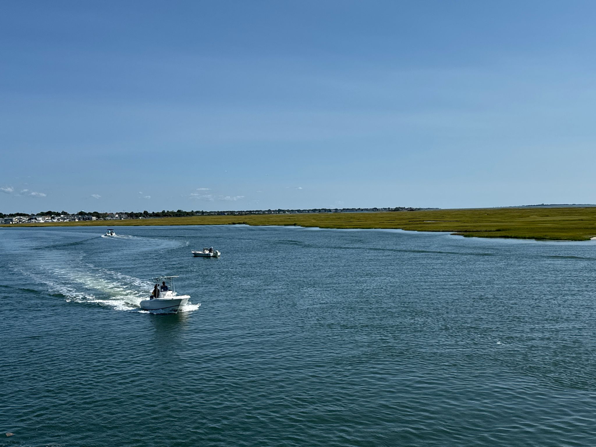 Several boats navigate a wide waterway bordered by green marshlands under blue sky. - Ocean Parkway Coastal Greenway