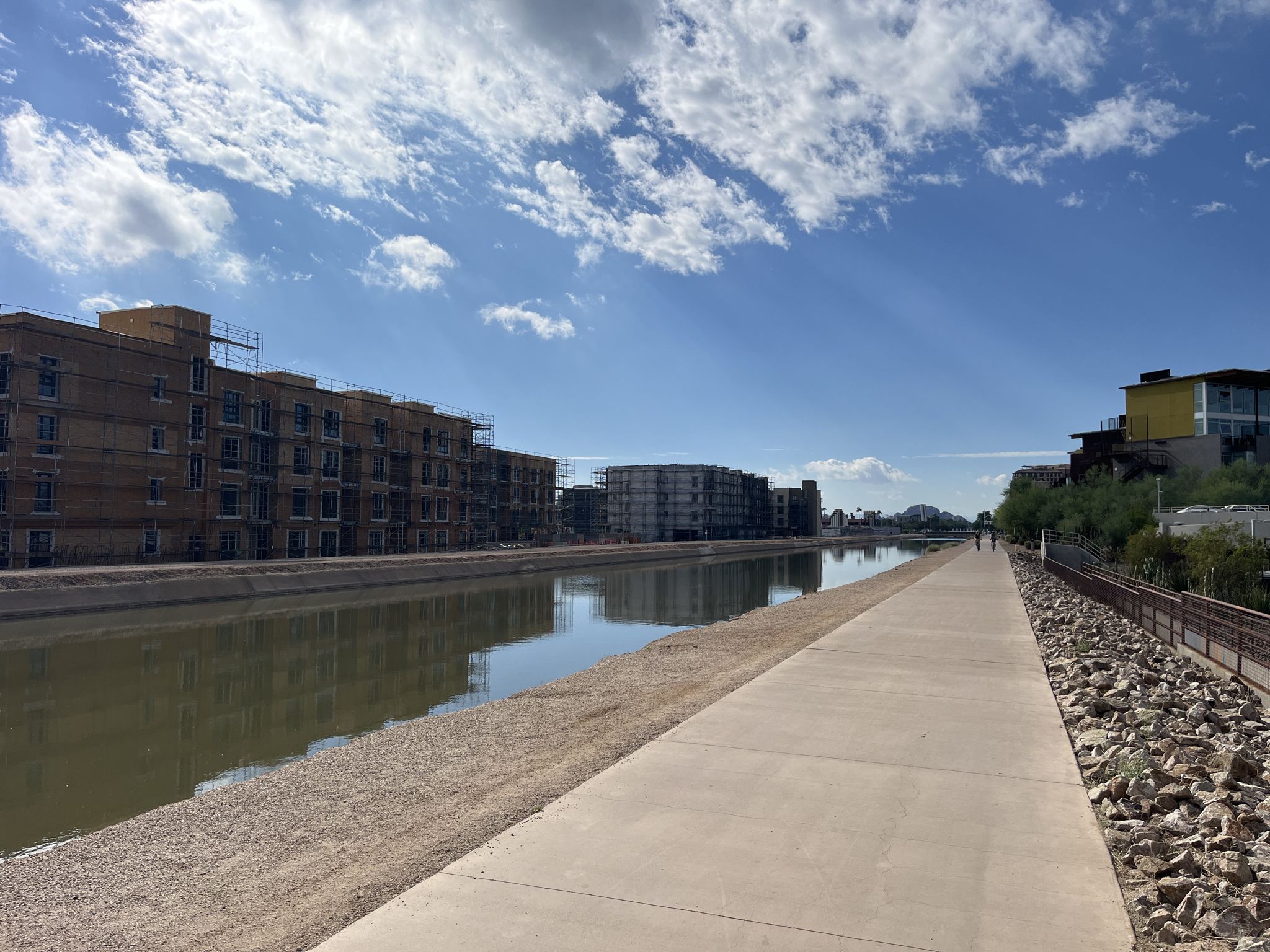 A canal with a paved path, buildings under construction, under a sunny sky. - Arizona Canal Trail