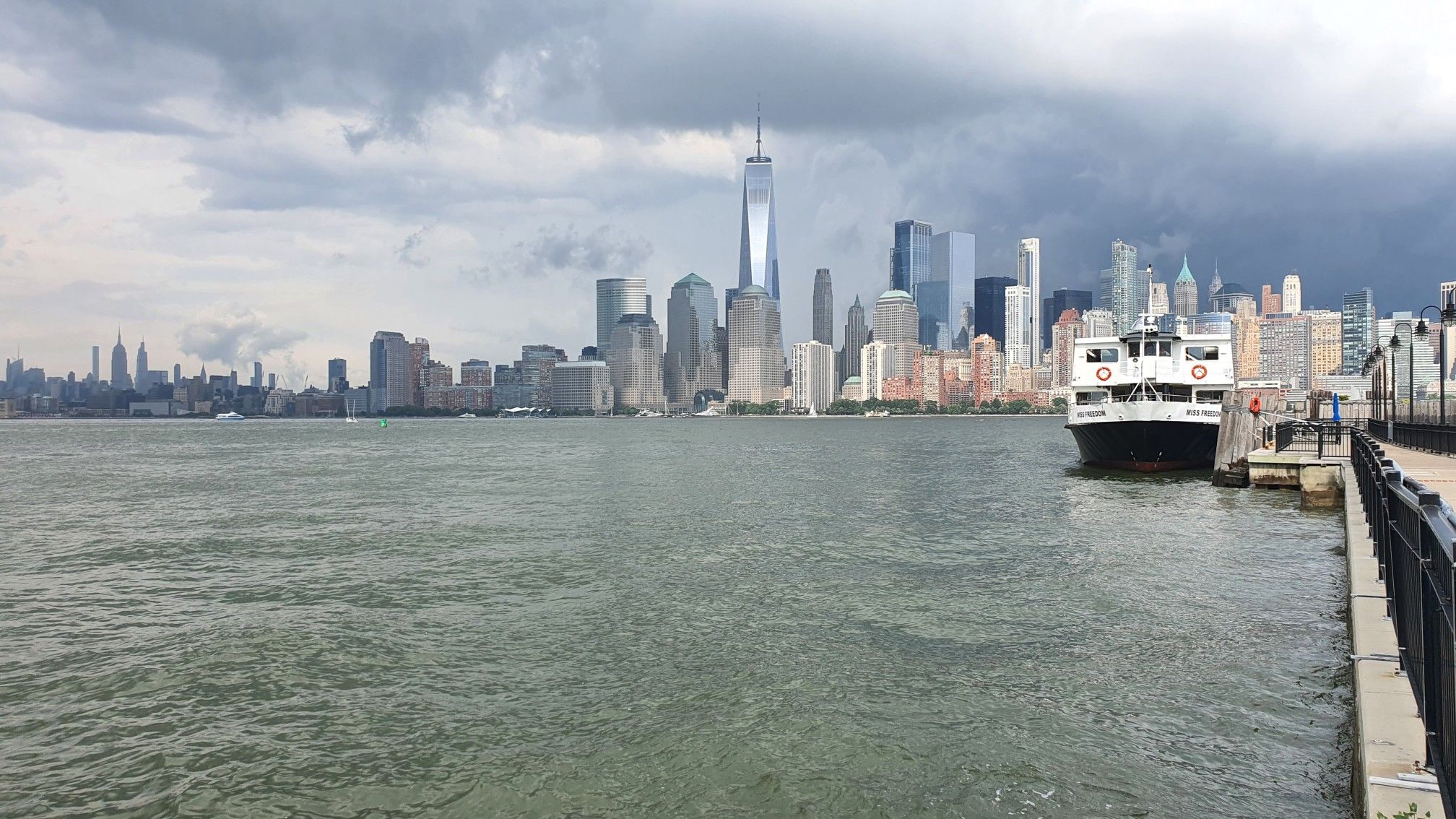 City skyline across choppy water with a docked ferry under stormy clouds. - Reclining Liberty – View of Ellis Island loop from Jersey City