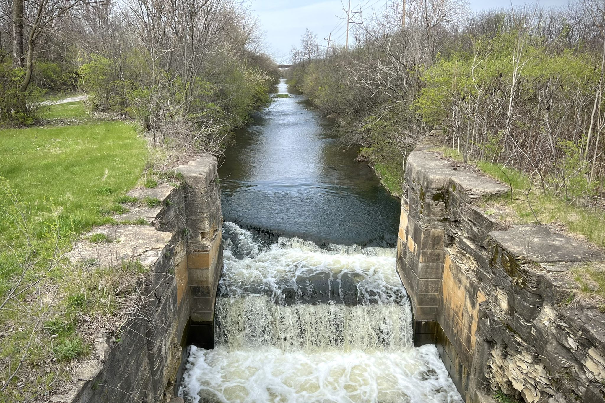 Water cascades over a small dam in a narrow canal with concrete walls. - Illinois and Michigan Canal Trail