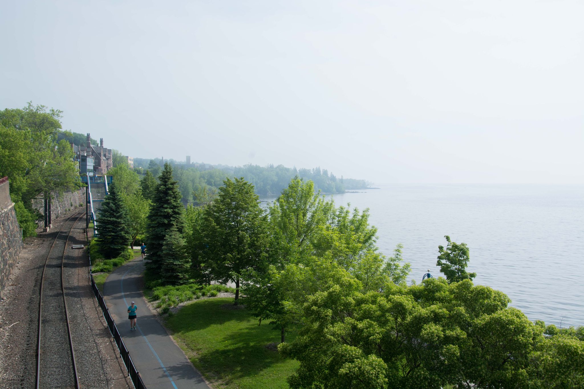 Person walks on a paved path next to railroad tracks and water. - Lakewalk loop from University of Minnesota Duluth