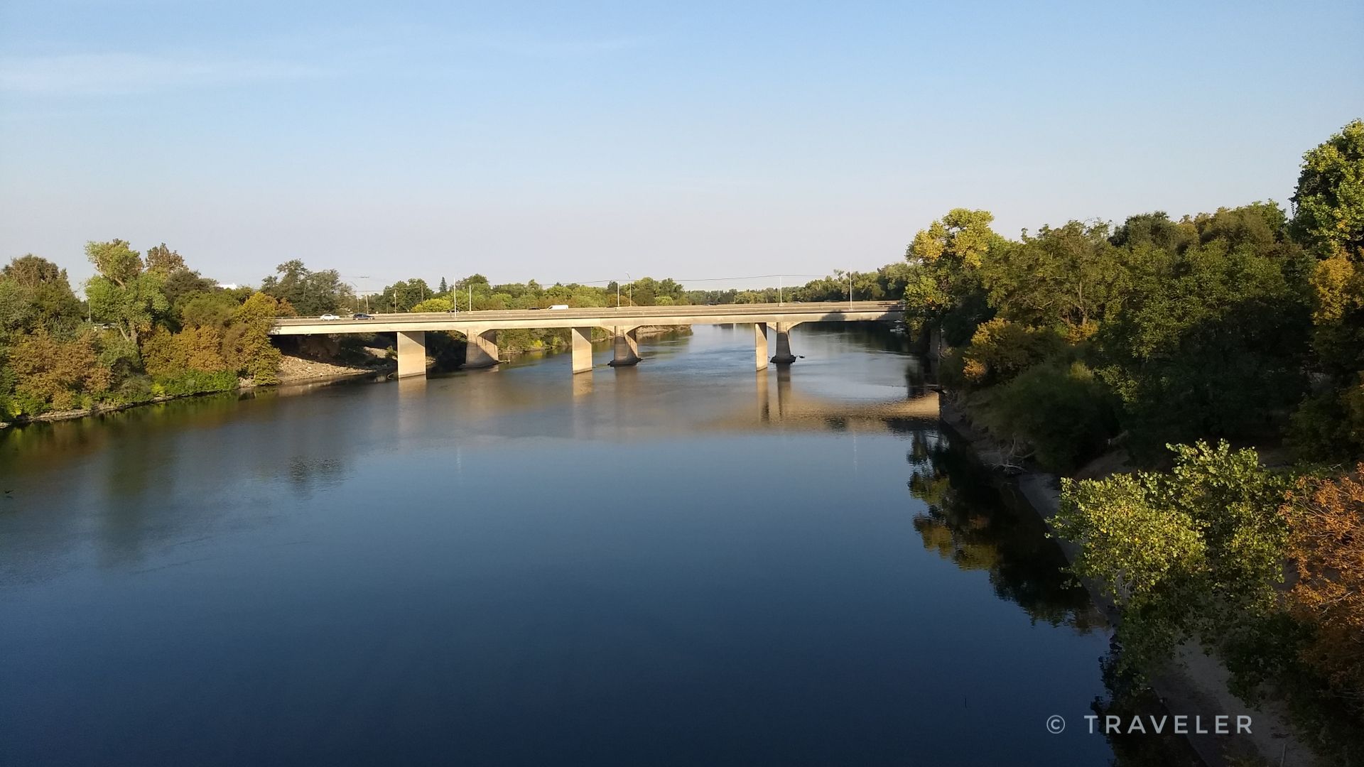 A concrete bridge spans a wide river with green trees under a clear sky. - American River and Sacramento River Trail Loop