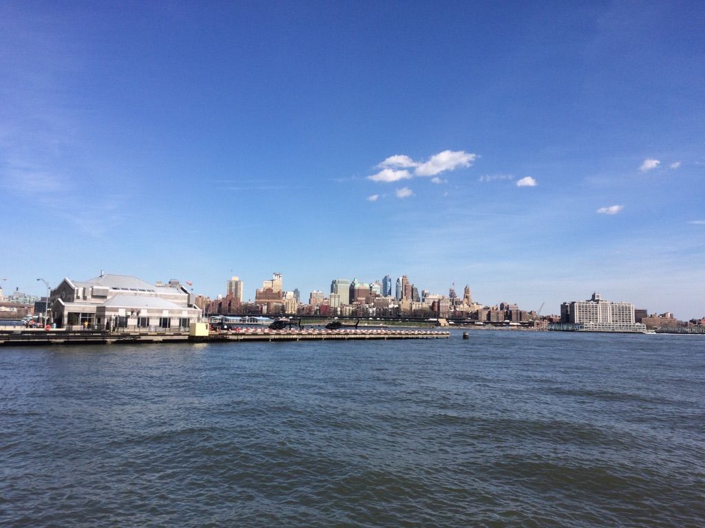 City skyline across a wide river with buildings and a clear blue sky. - One World Trade Center – East Coast Memorial loop from Wall Street