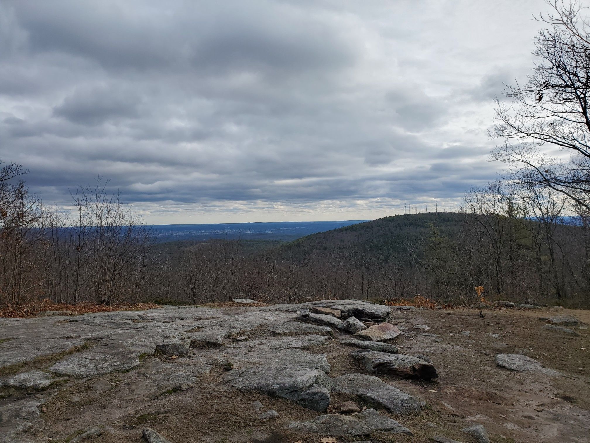 Rocky overlook with bare trees and distant hills under a cloudy, grey sky. - North Uncanoonuc Mountain via White Dot and Red Dot Trail