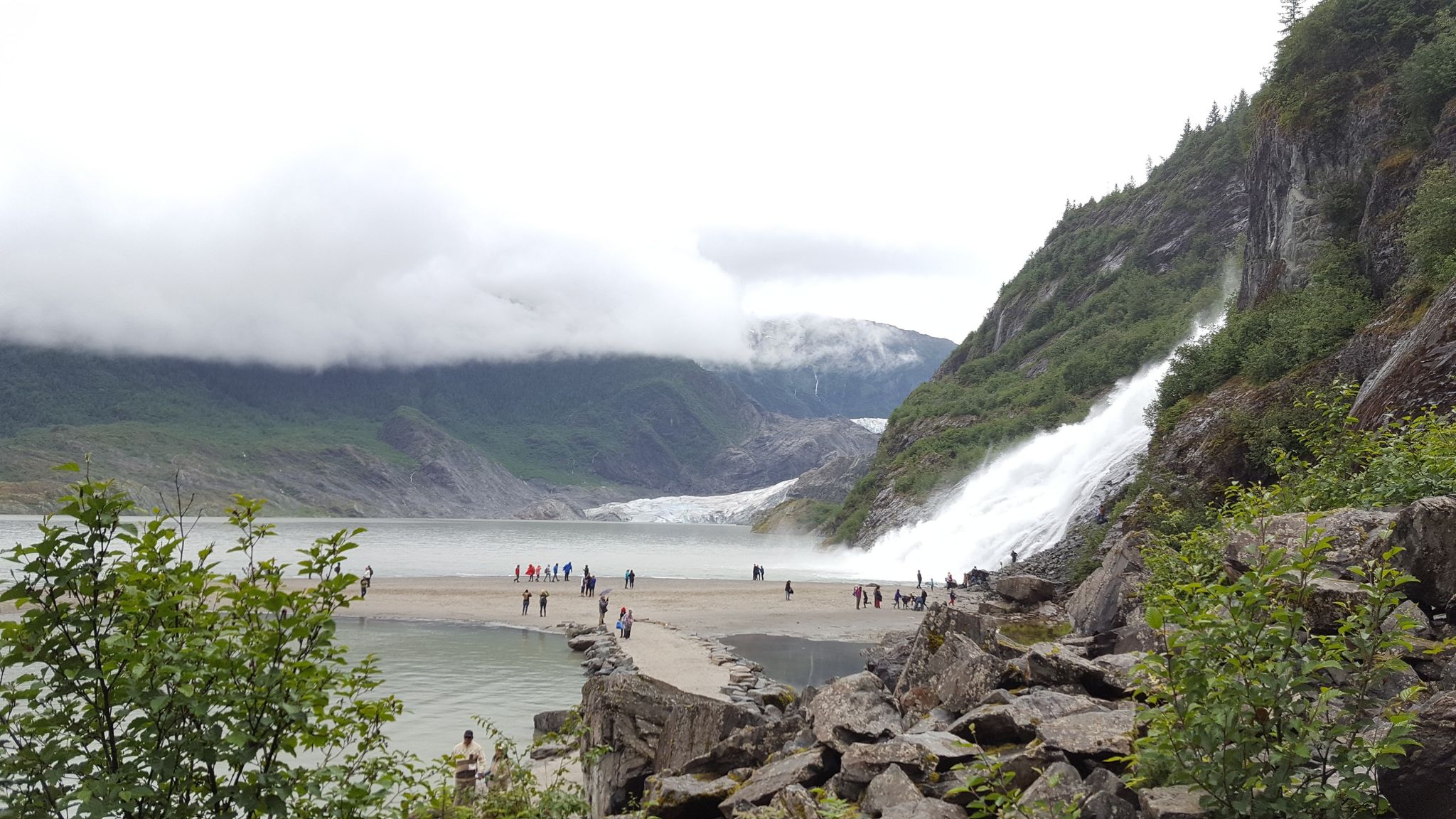 People gather near a powerful waterfall cascading into a lake with a distant glacier. - Nugget Falls Trail