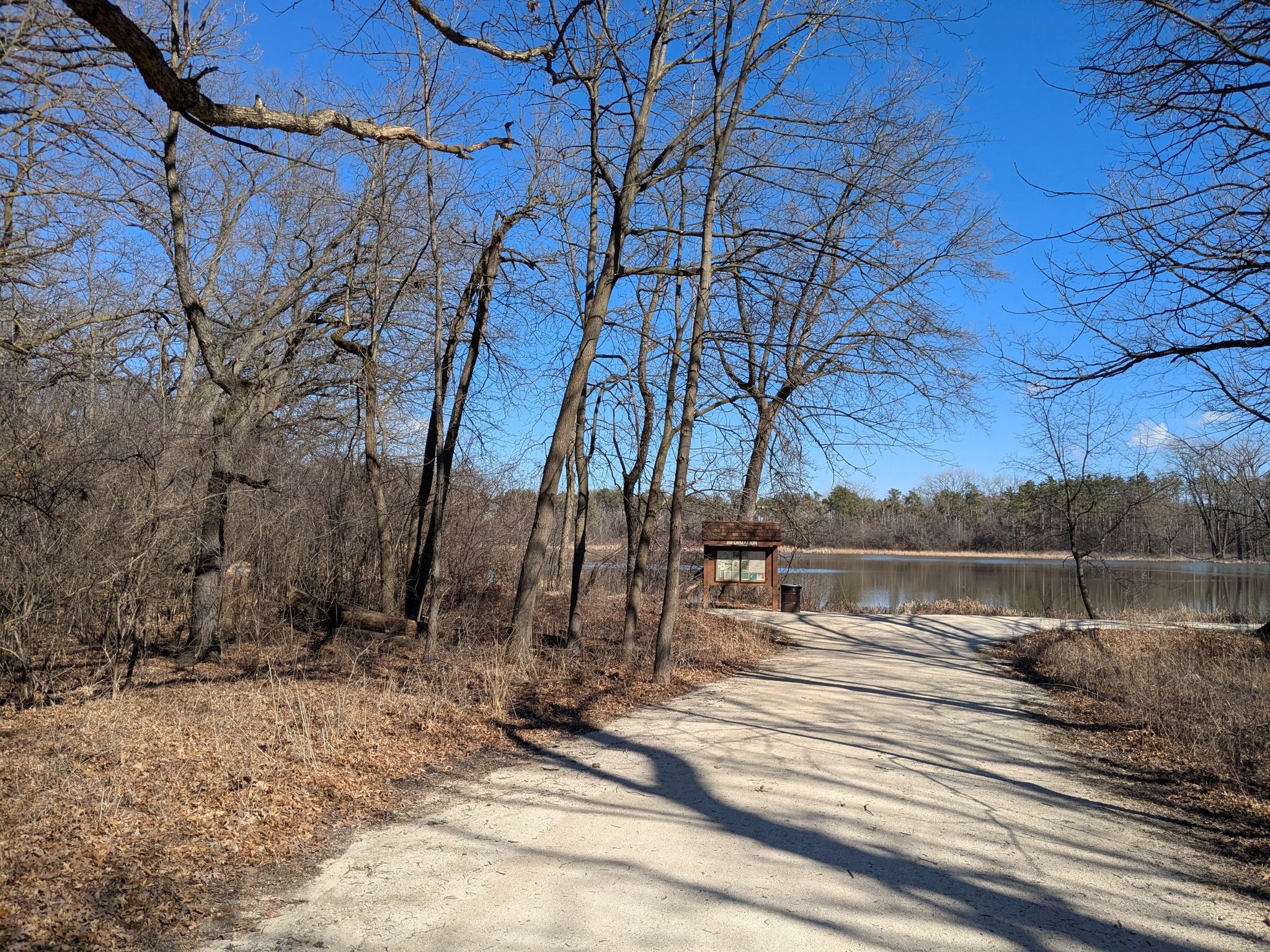 Dirt path leading to a lake with a wooden sign, surrounded by bare trees. - Waterfall Glen Main Trail