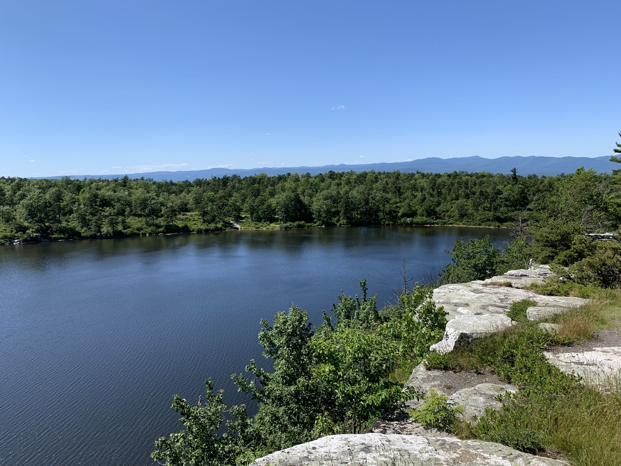 Sunny view of a calm lake surrounded by dense green forest and distant mountains. - Lake Minnewaska – Lake Awosting viewpoint loop from Rainbow Falls