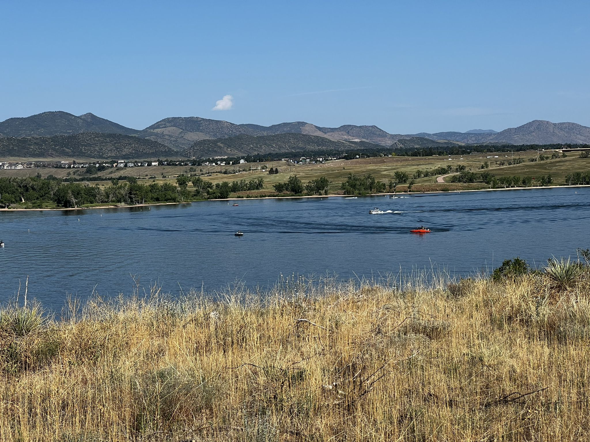 A wide lake with boats, dry grass in foreground, and distant mountains under blue sky. - Chatfield Internal Trail