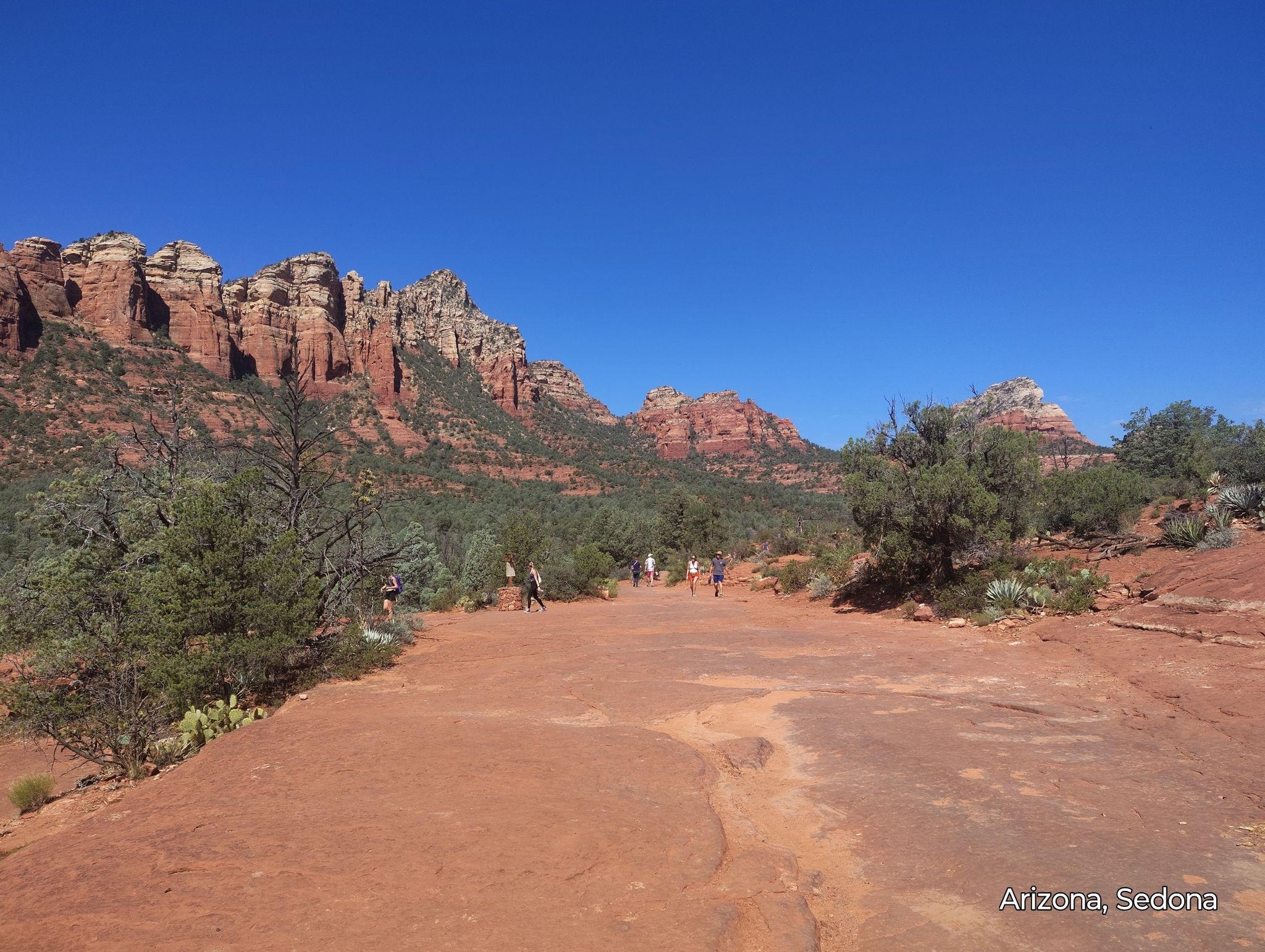 Hikers walk on a wide red dirt path with distant red rock mountains. - Grand Central, Ant Hill, and Adobe Jack Loop