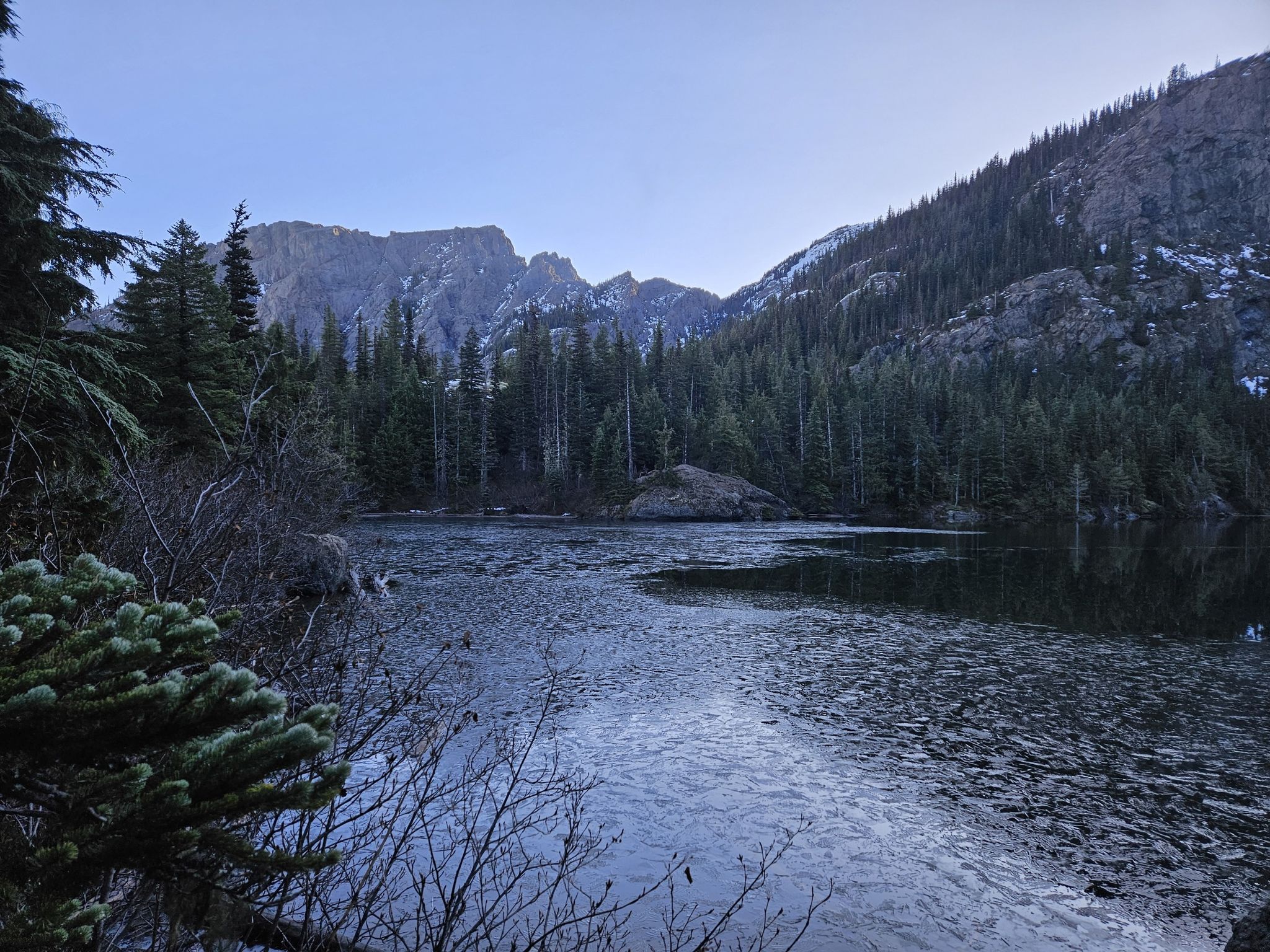 Icy lake surrounded by dark evergreen forests and mountains under a pale sky. - Lake Angeles Trail
