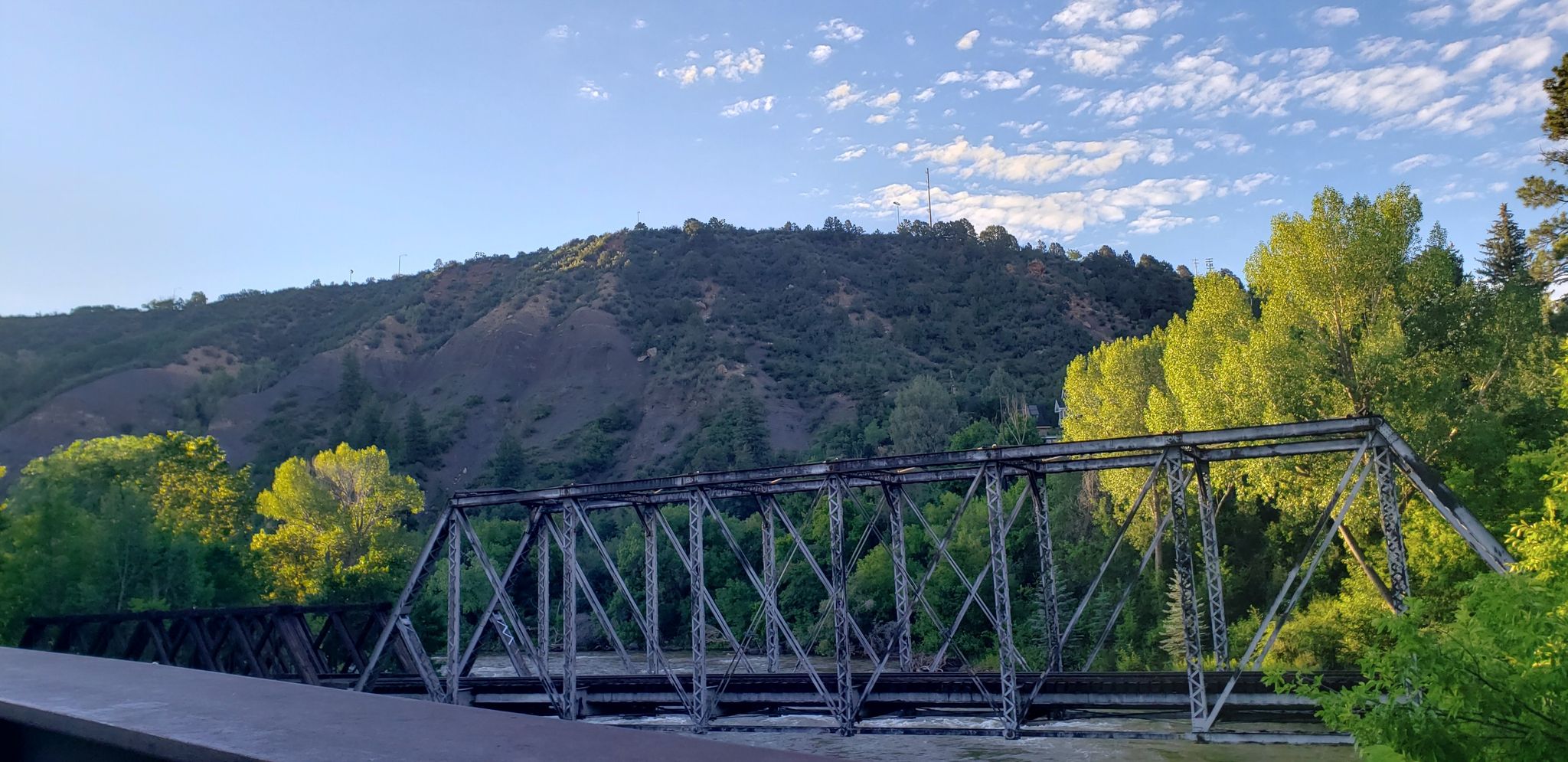 A metal truss bridge crosses a river with sunlit trees and a large hill. - Animas River Trail