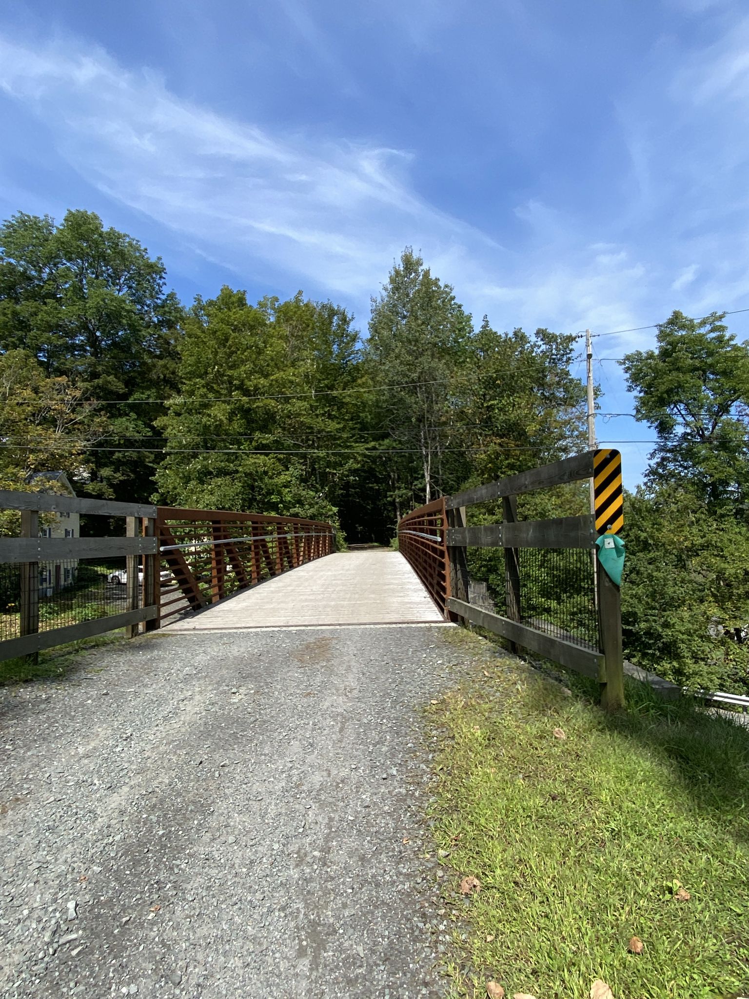 A metal bridge over a gravel path, surrounded by green trees, under blue sky. - Wonderful Lamoille Rail Trail – Public Access Tools and Pump loop from Saint Johnsbury