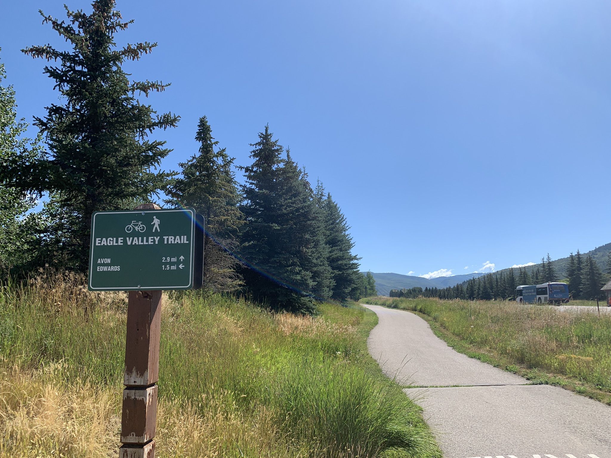 A paved path next to a trail sign, pine trees, and distant mountains. - Market Square at Beaver Creek – Nottingham Lake loop from Beaver Creek