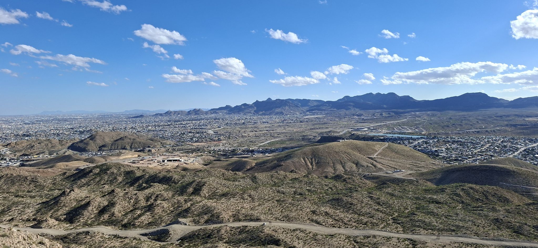Wide view of desert hills, a city, and distant mountains under a cloudy sky. - Mount Cristo Rey Trail