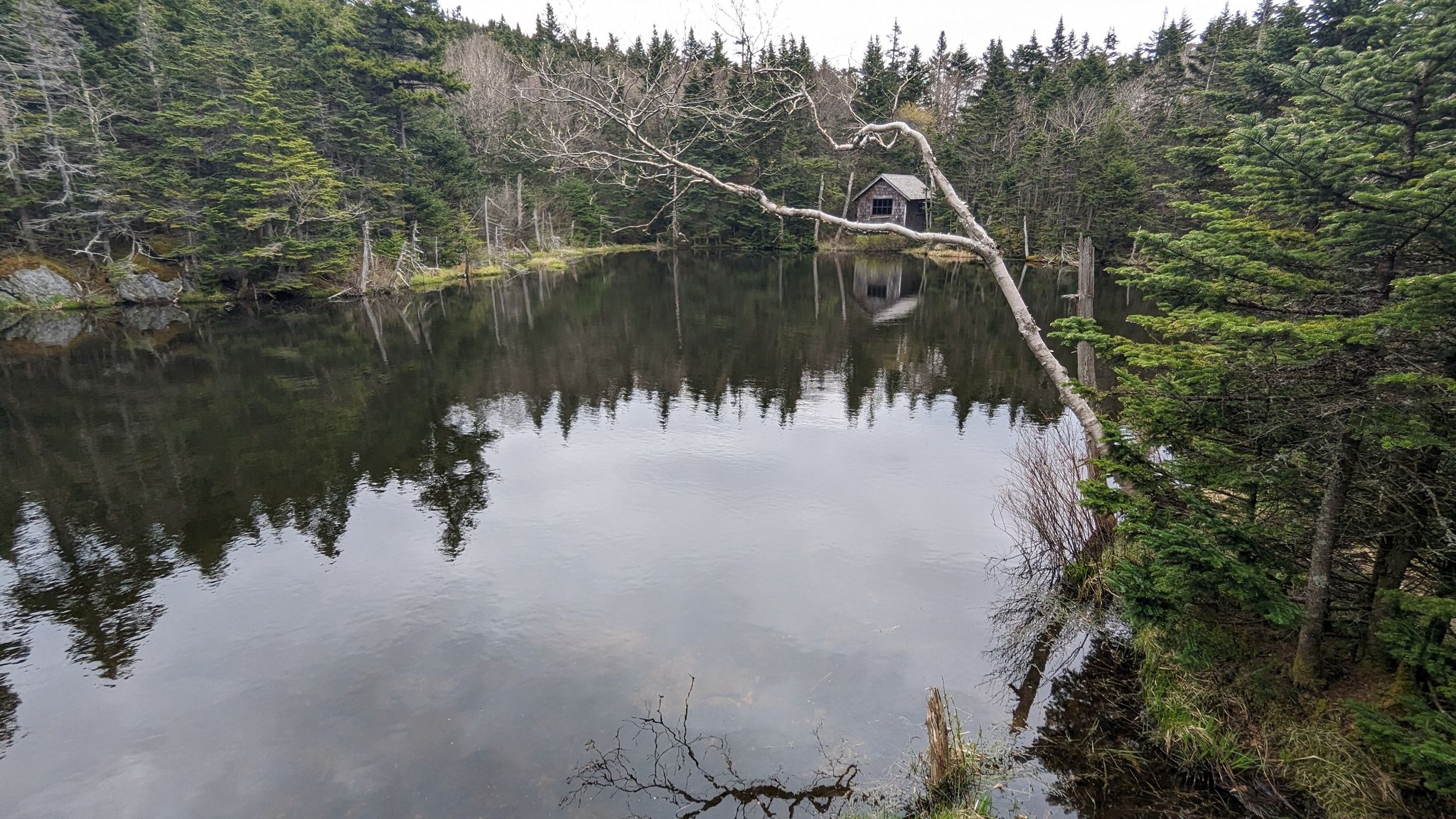 A small cabin reflected in a dark, still lake under an overcast sky. - Cheshire Harbor Trail