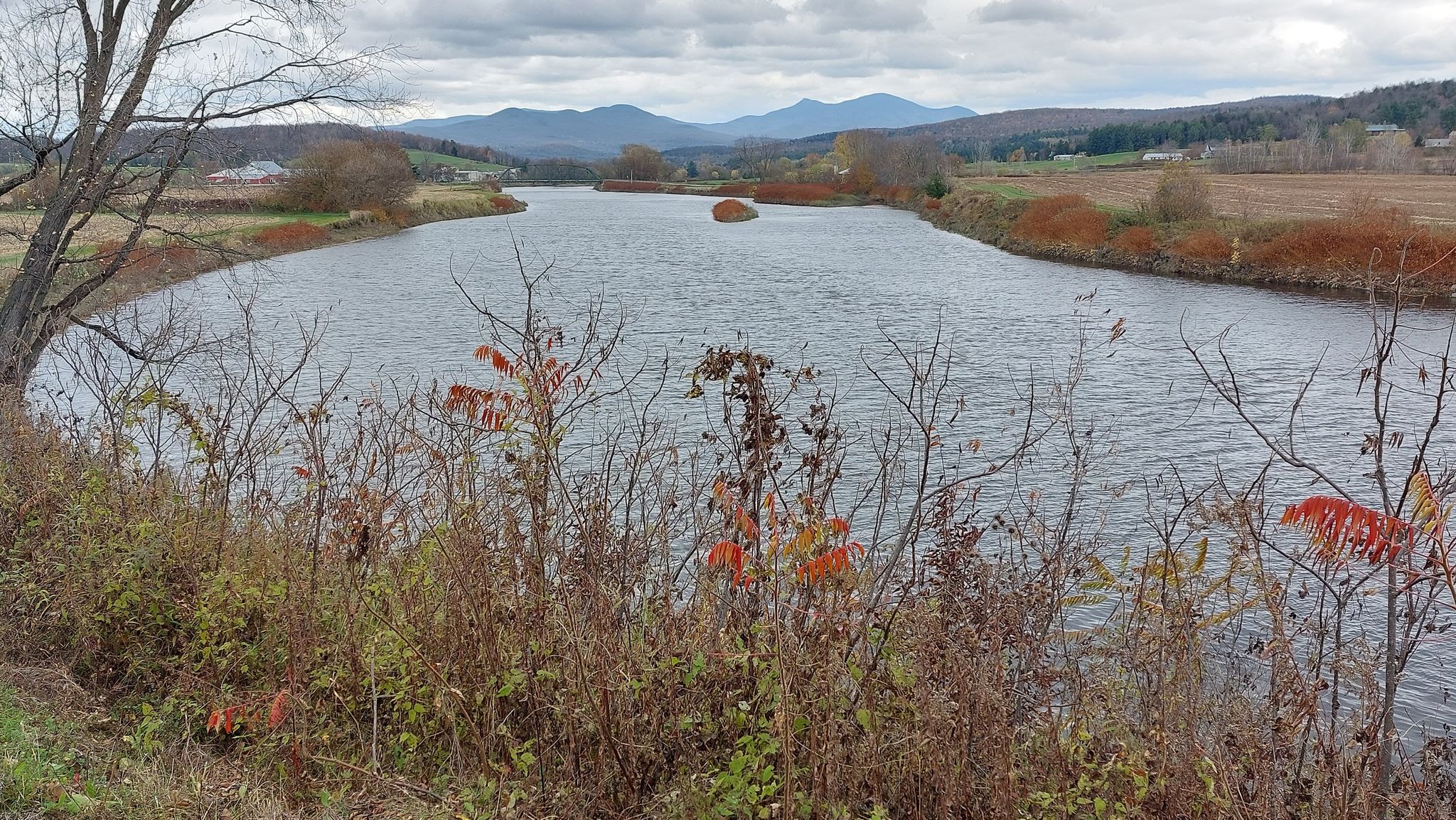Wide river with autumn-colored banks, fields, and distant mountains under cloudy sky. - View of the Missisquoi River – Missisquoi River loop from Richford
