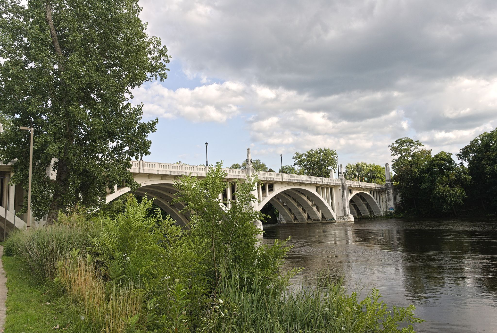 A large, arched concrete bridge spans a river with lush green banks. - St Joseph River bridge loop from South Bend