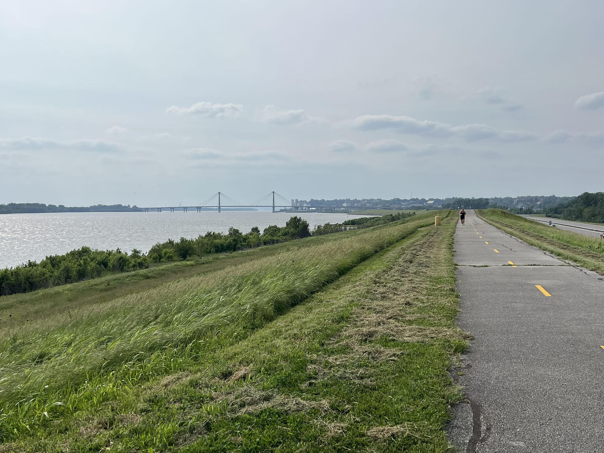 A person walks on a paved path next to a wide river under an overcast sky. - MCT Confluence Trail
